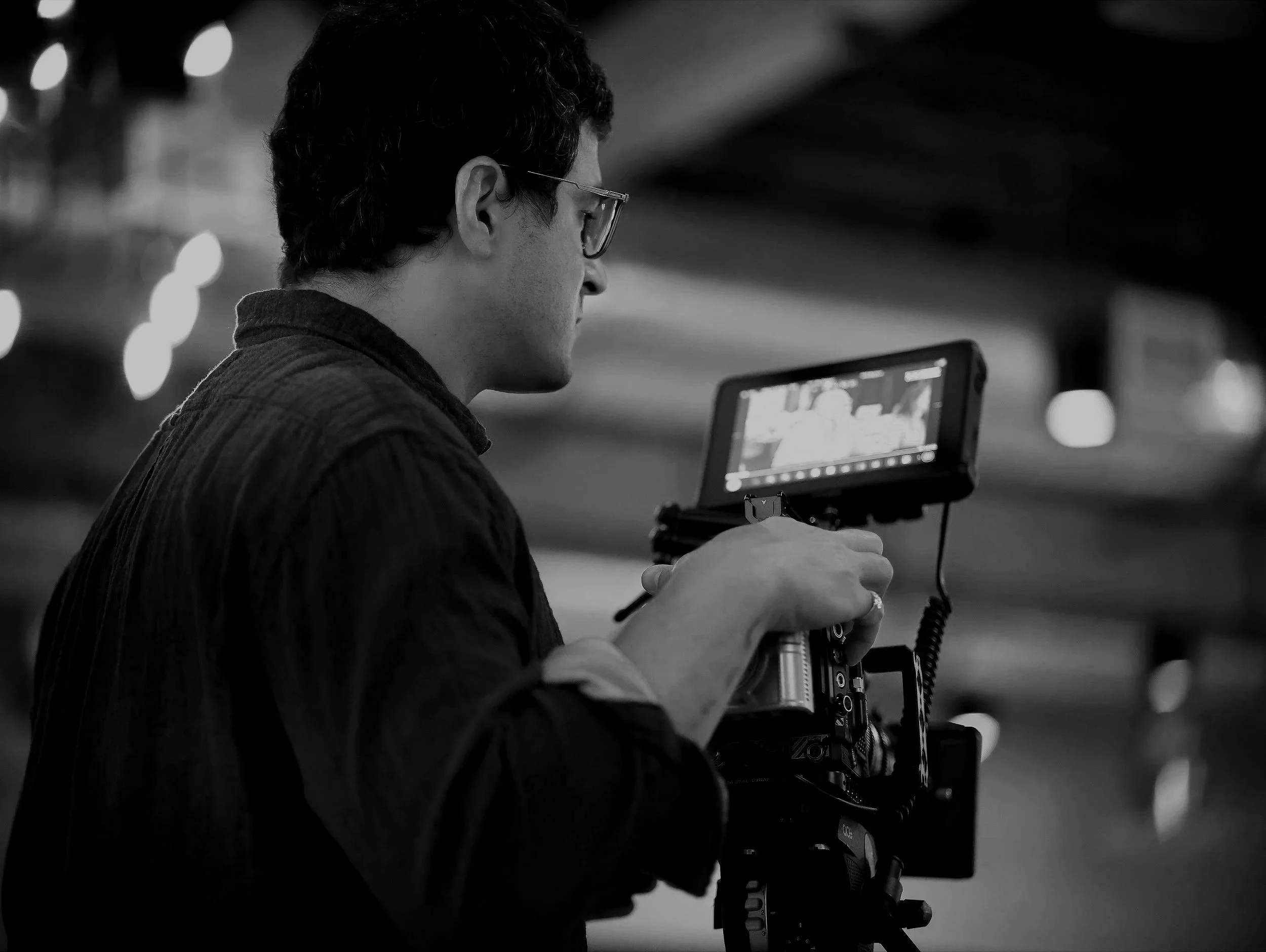 A man with short dark hair and glasses operating a camera monitor in a dimly lit indoor environment.