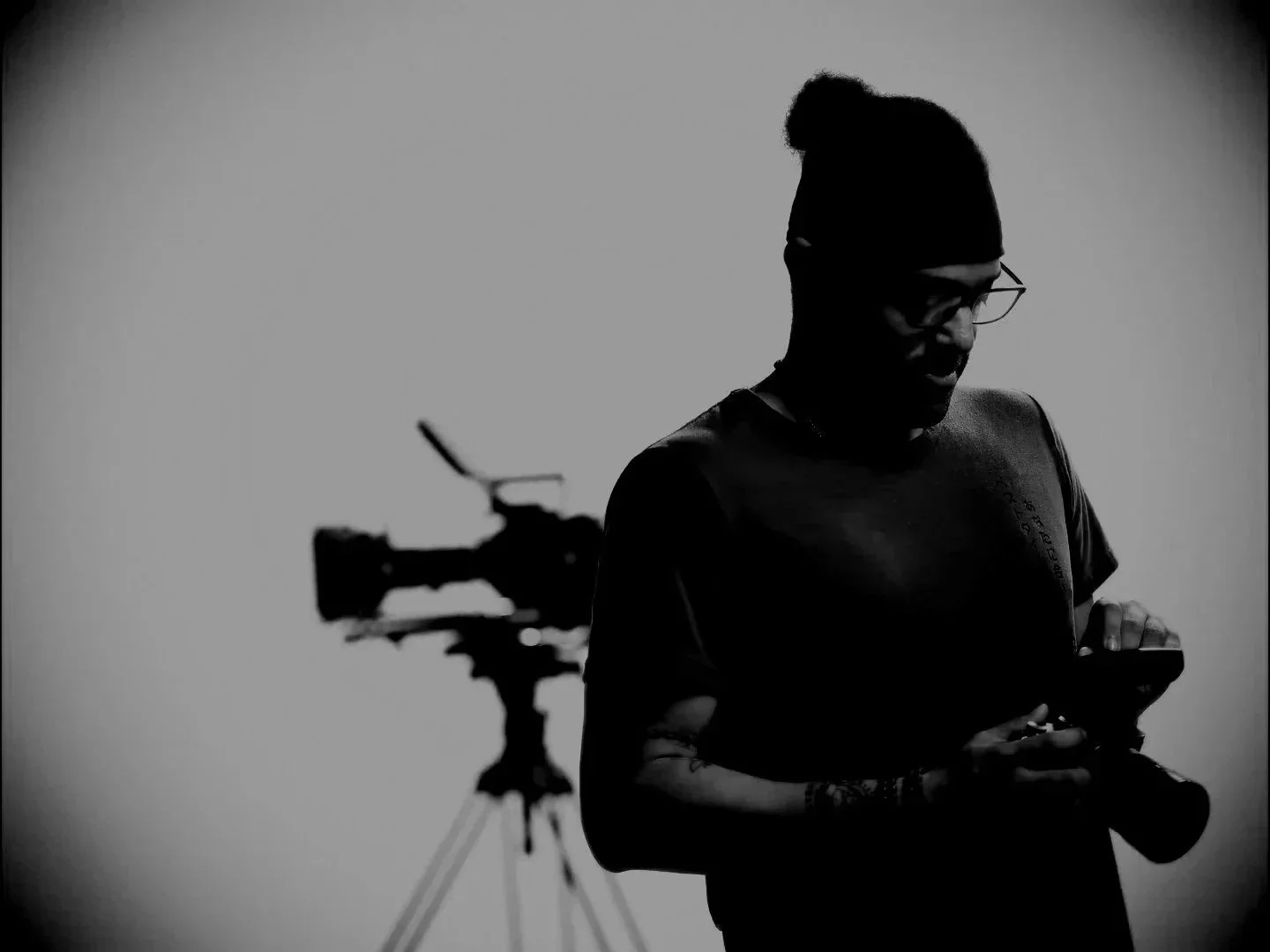 Silhouette of a person wearing glasses and a beanie, holding a smartphone, with a camera on a tripod in the background against a plain wall.