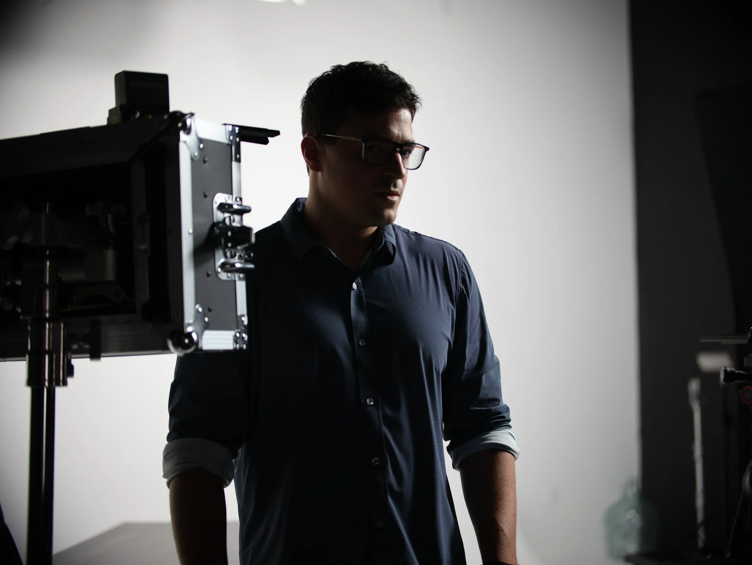 A man with dark, curly hair, wearing glasses and a dark blue dress shirt, stands in a photography studio with a neutral background and professional lighting equipment.