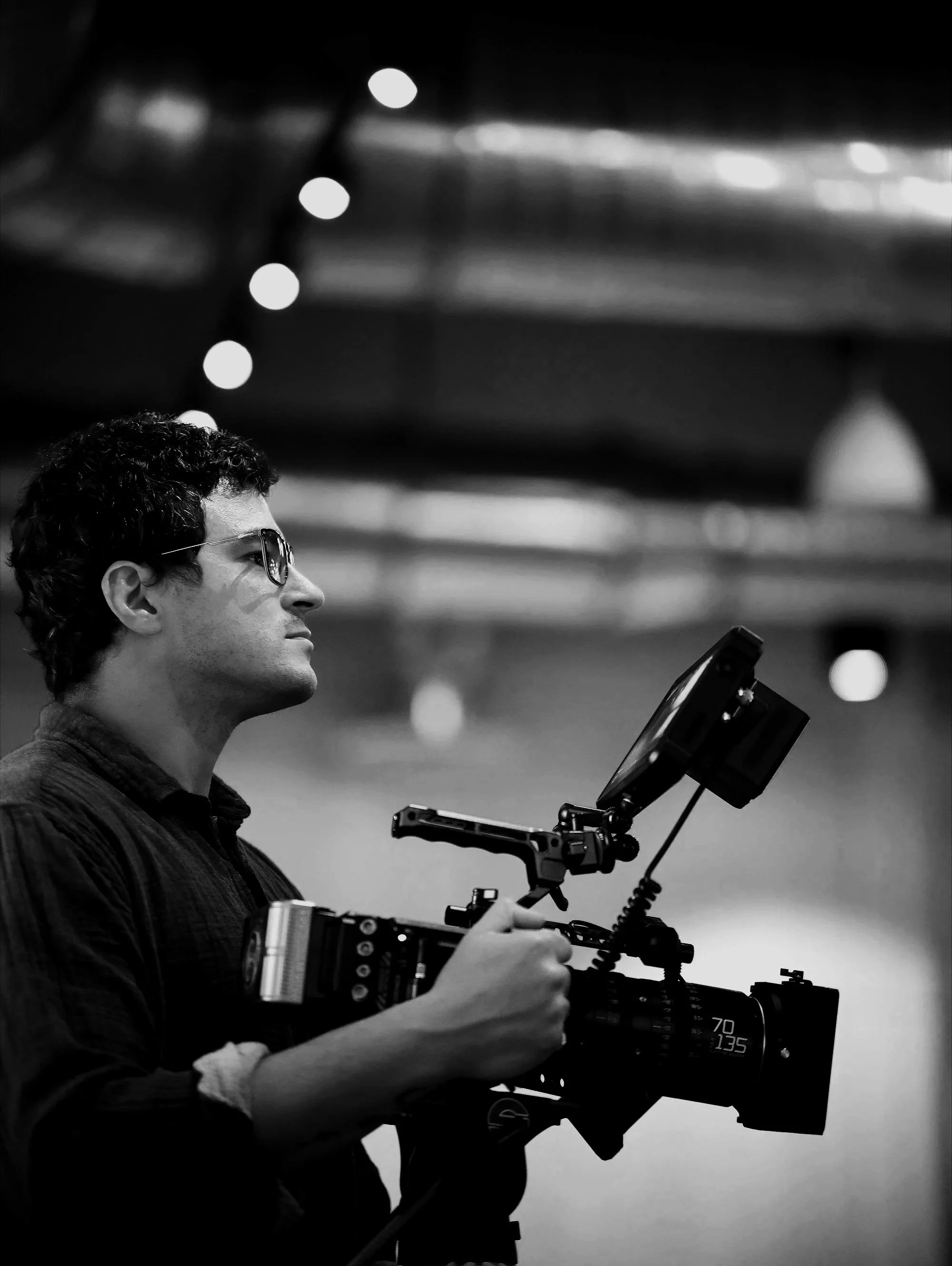 A man operating a professional camera, wearing glasses, in a low-light environment with blurred lights in the background.