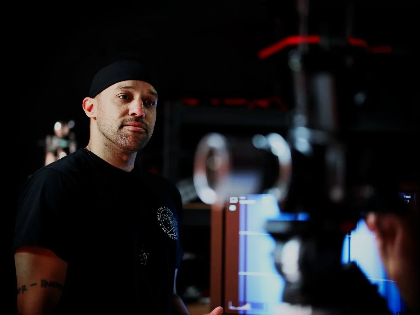A man with a black beanie and black shirt standing indoors, looking at the camera. There are blurred objects and equipment in the background.
