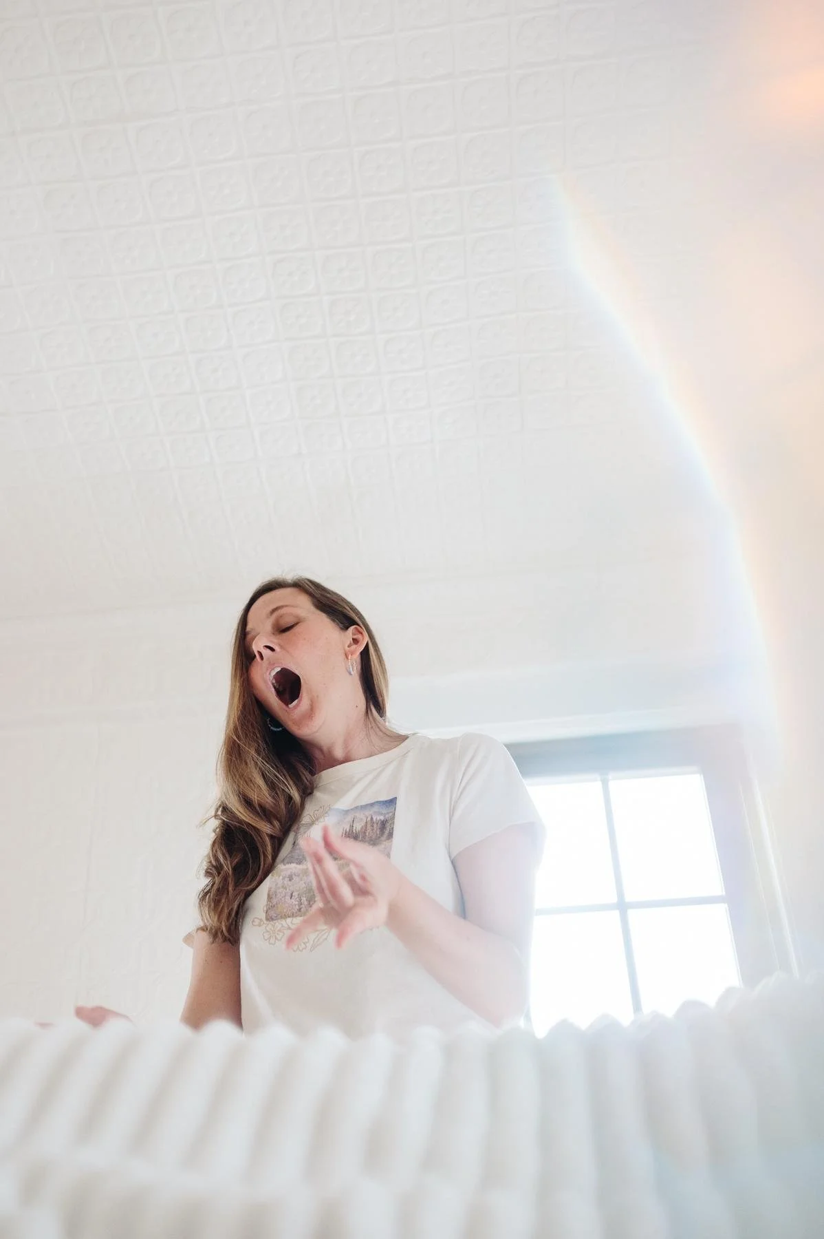 Woman with mouth open with her hand lifted beside a window as a soft rainbow of light falls across the wall, expressive branding portrait, energy worker
