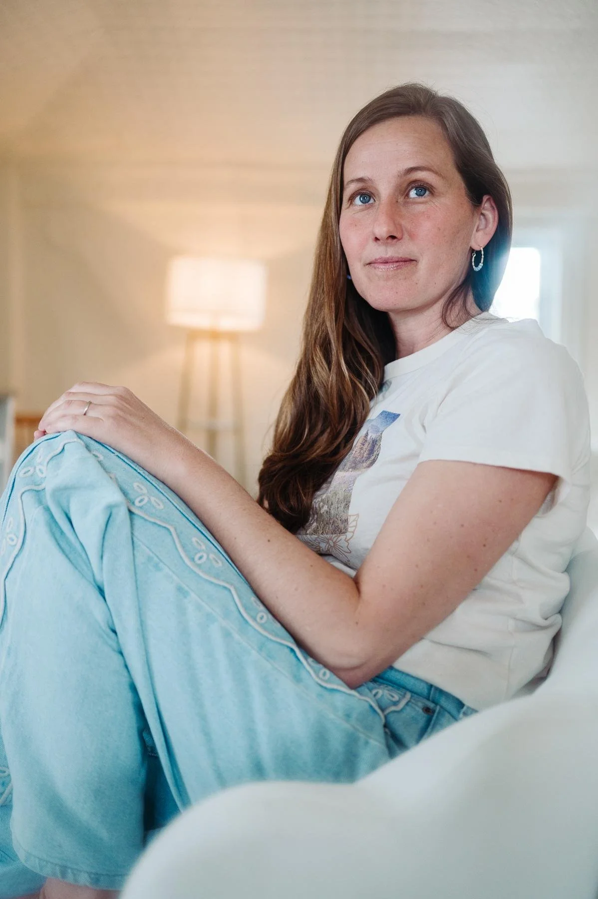 Woman in a white tee and light jeans sitting on a white chair in warm window light, North Andover branding photographer
