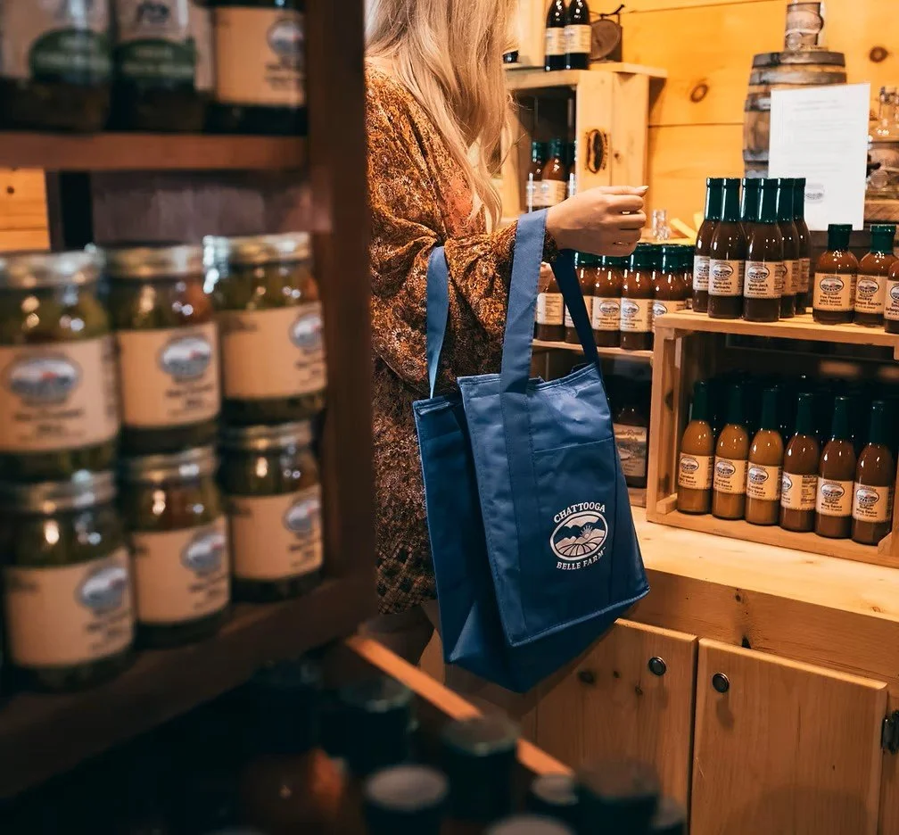 A woman shopping at a farm store, holding a blue tote bag with a mountain logo and the words 'Chattoga Belle Farm,' surrounded by shelves of jars and bottles.