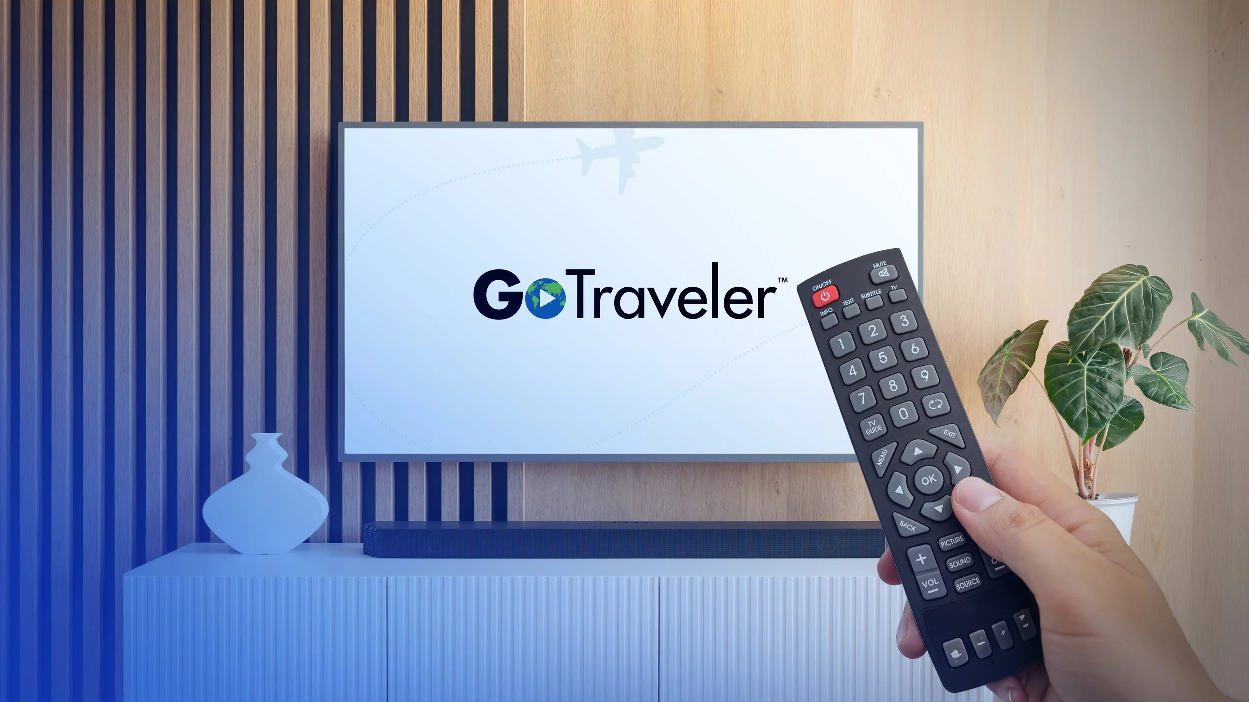 A person holding a TV remote pointing at a flat-screen television displaying the Go Travel logo with a stylized globe and airplane graphic. The TV is on a white modern entertainment stand with a decorative vase and a large potted plant nearby.