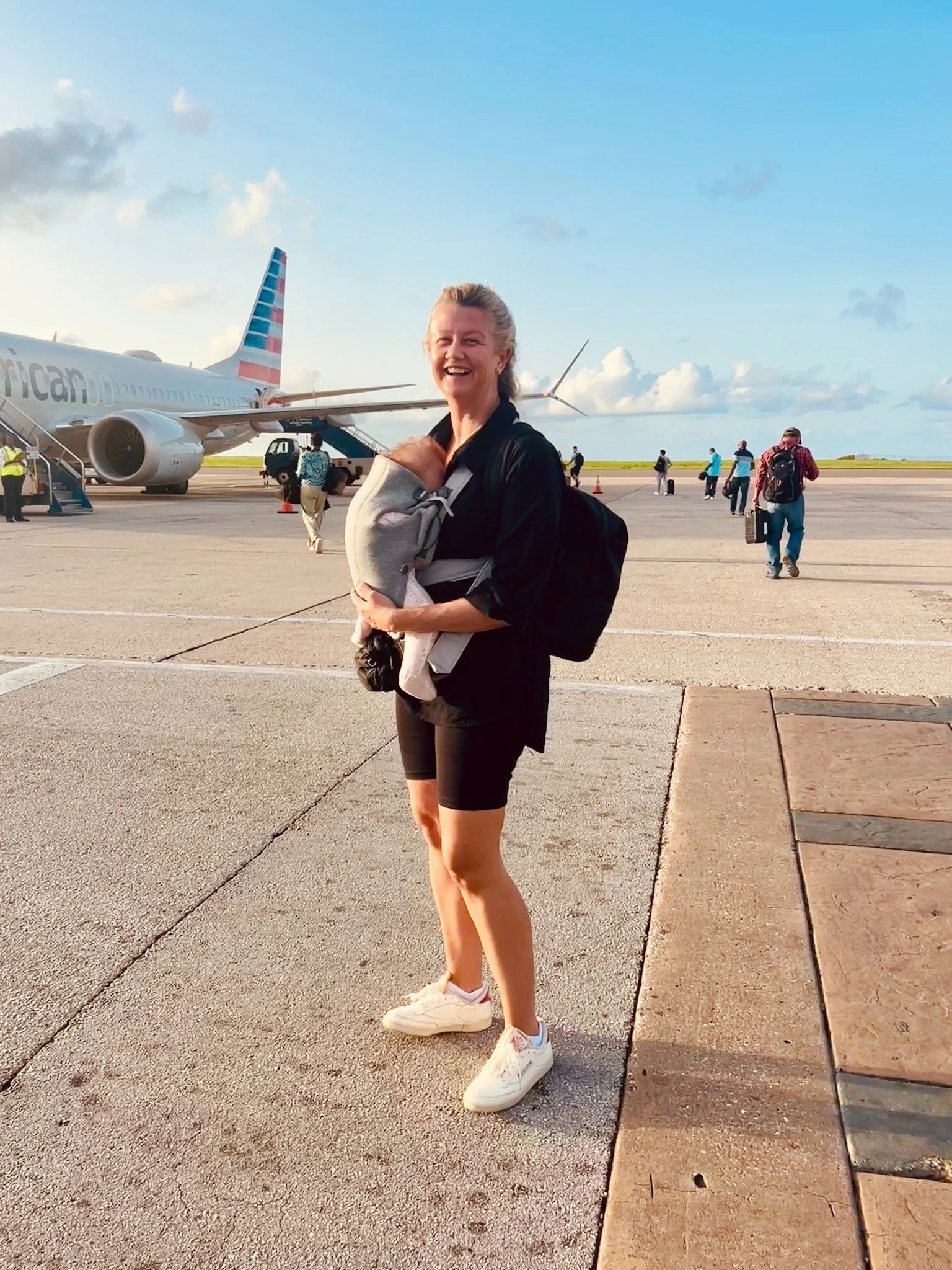 Malou Morgan standing on the tarmac in front of an American Airlines plane, smiling while holding her baby in a carrier at sunrise.