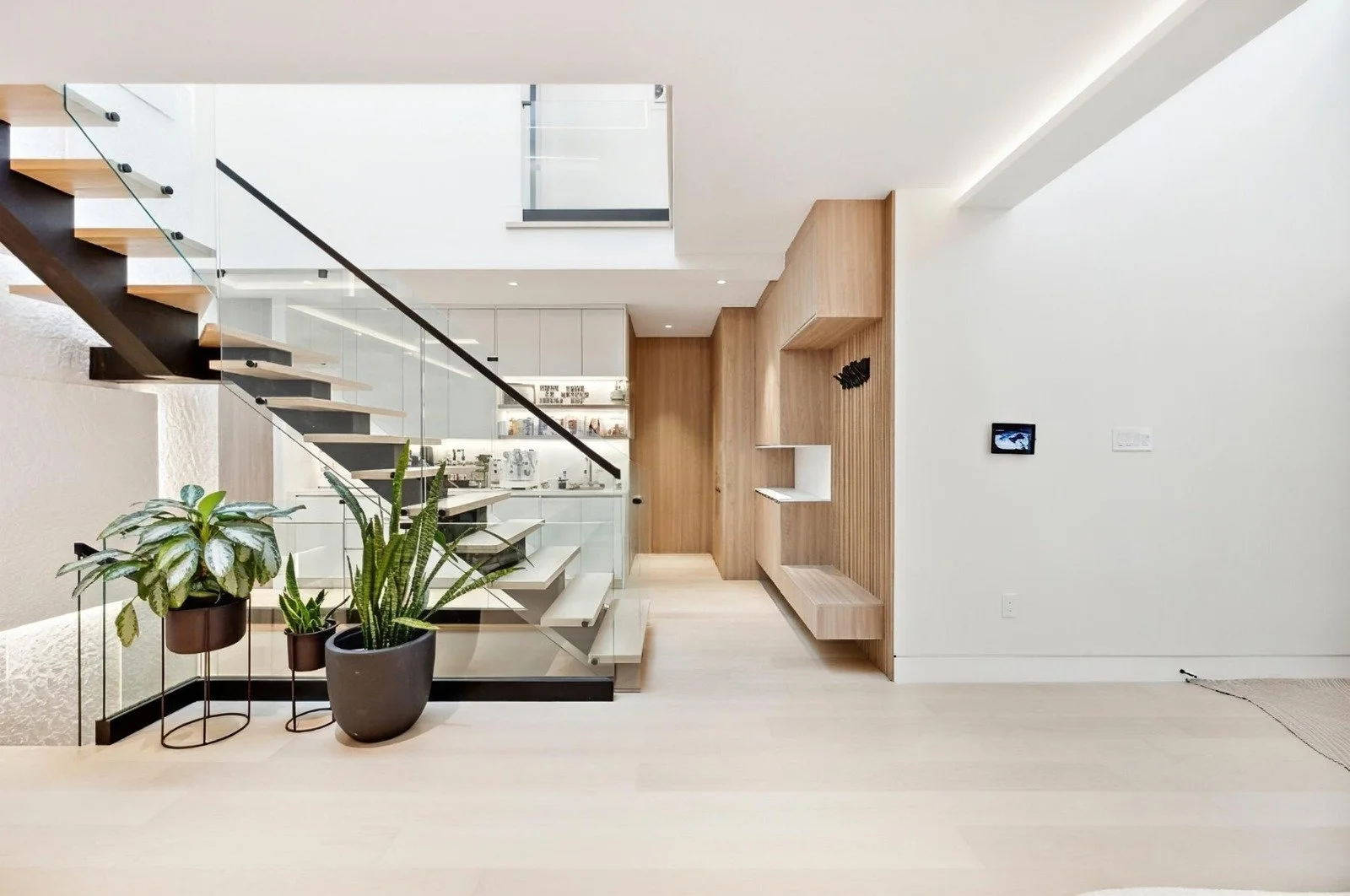 Modern interior with wooden staircase and plants near glass railing, kitchen area in background, white walls, and minimal decor.