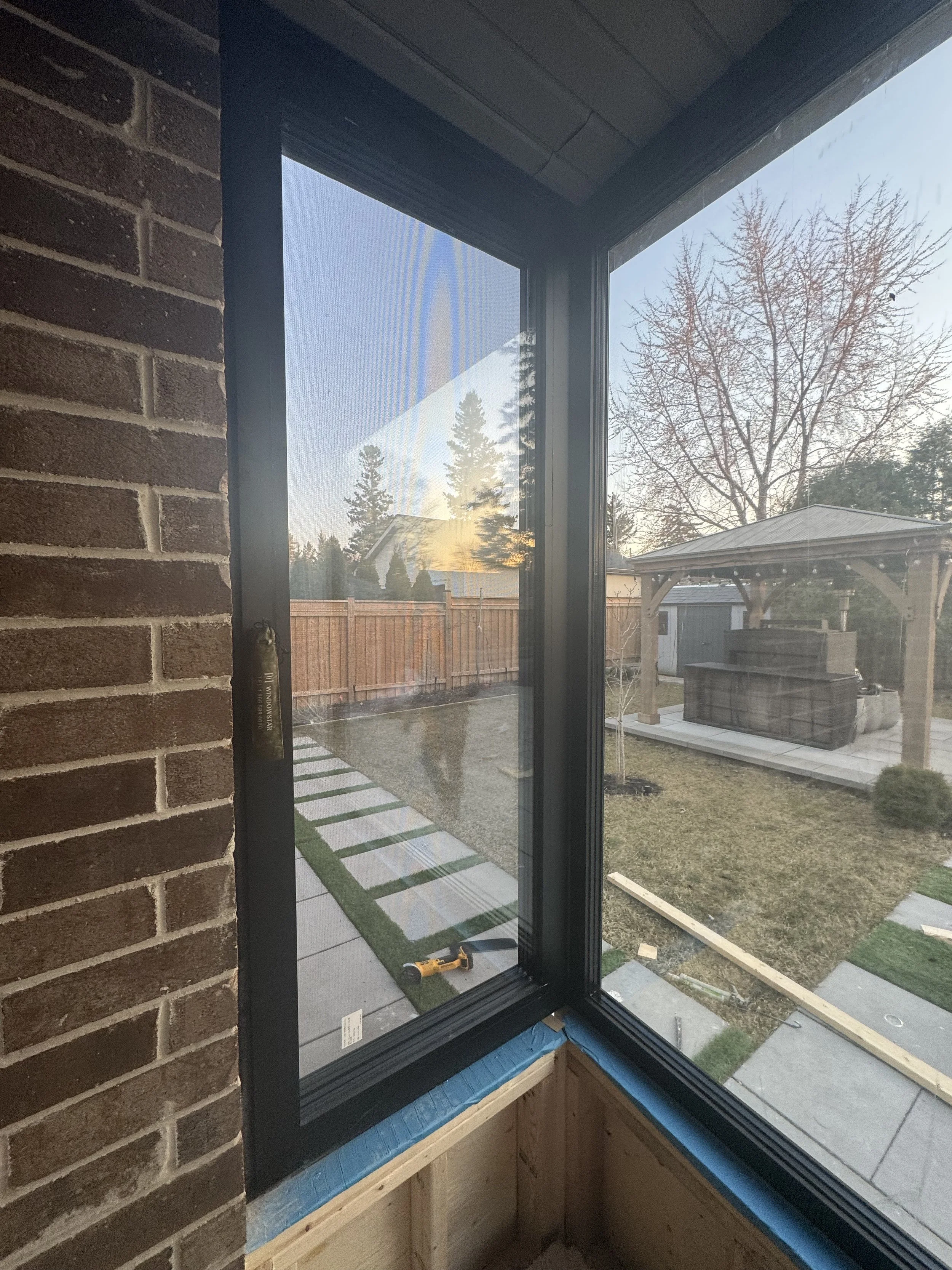 View from inside a house under renovation, looking out a window. The scene outside includes a backyard with a wooden fence, trees, a small shed, and a covered outdoor structure. The interior has a brick wall and construction materials on the windowsill, with tools visible.