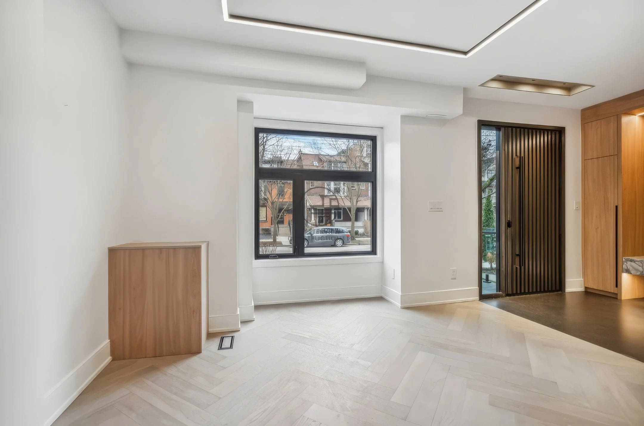 Bright modern living room with herringbone wood flooring, large window, recessed ceiling lighting, and open entryway with wood accents.