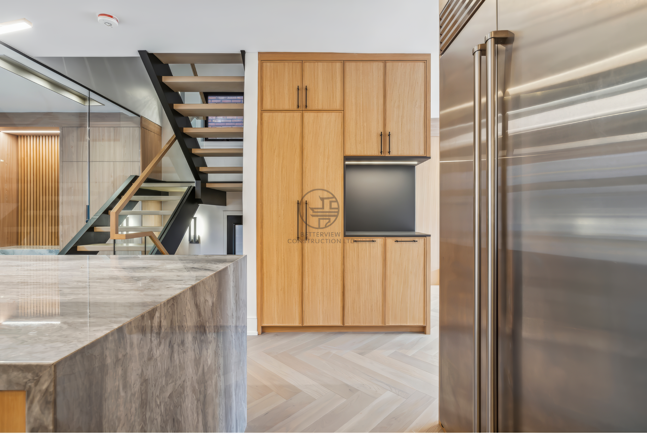 Modern kitchen with wood cabinetry, marble island countertop, stainless steel refrigerator, and open staircase in contemporary home design.