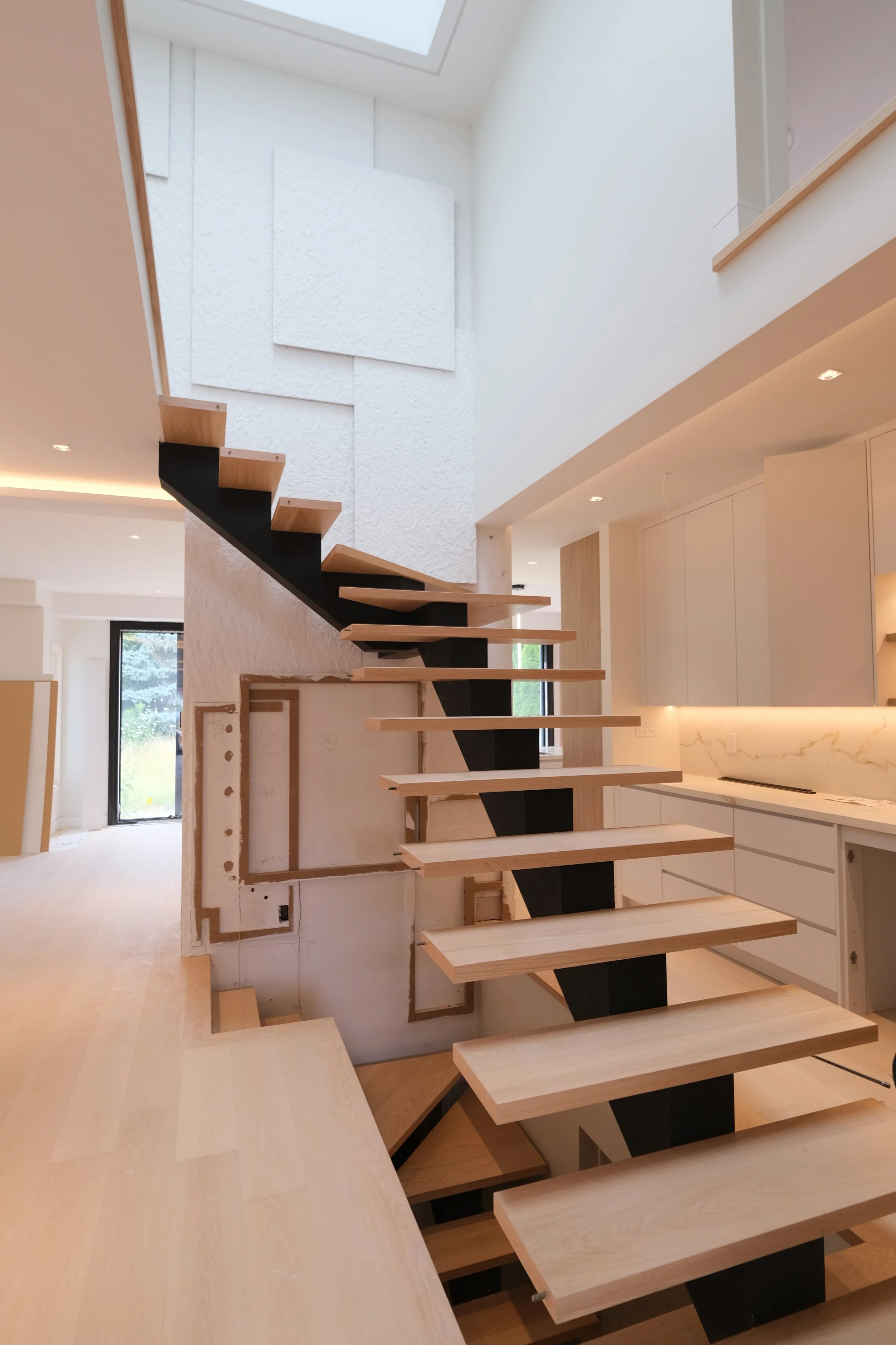 Interior of a modern home under construction with a floating staircase, white textured wall panels, and white cabinetry.