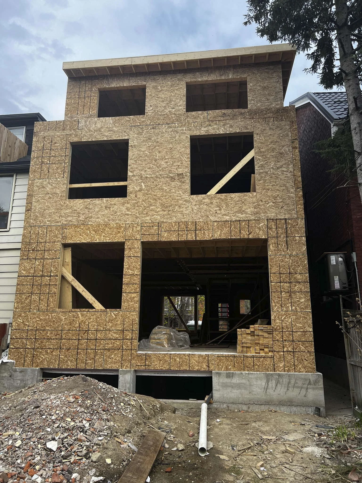 The front of a three-story house under construction with wood framing and plywood sheathing, open window spaces, construction debris, and piles of wood and gravel in the foreground.