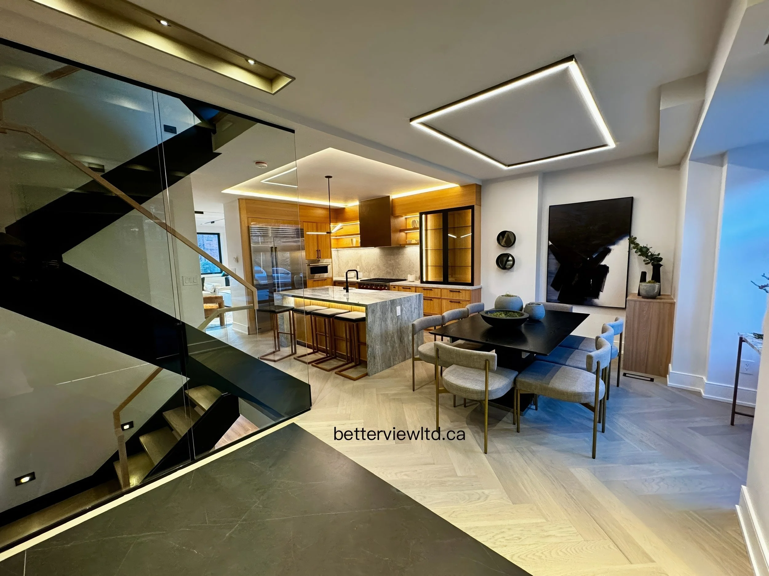 Modern open-concept kitchen and dining area with marble island, wood cabinetry, LED lighting, and floating staircase with glass railing.