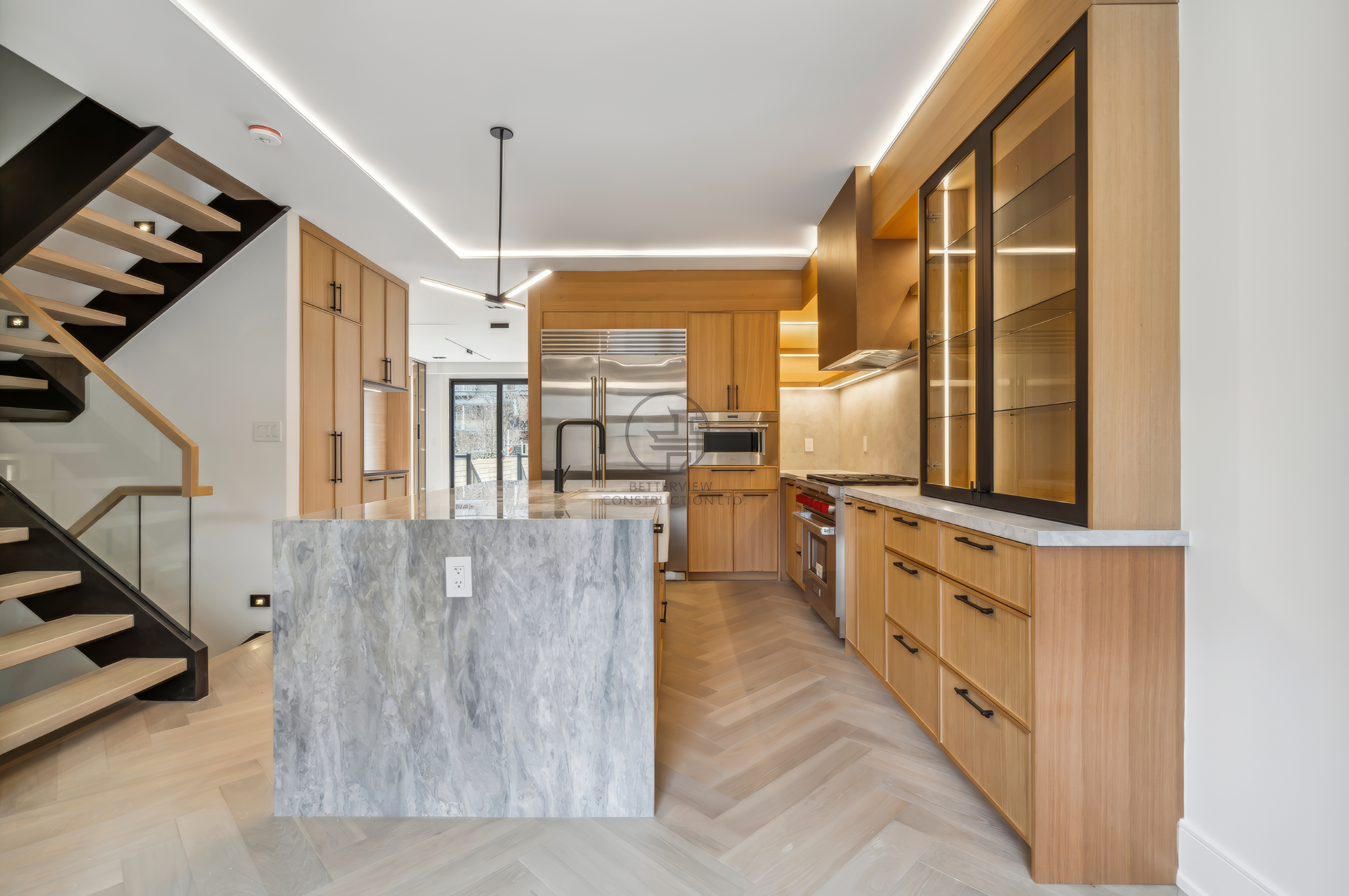 Open-concept modern kitchen with marble island, wood cabinetry, LED ceiling lighting, and adjacent floating staircase in contemporary home design.