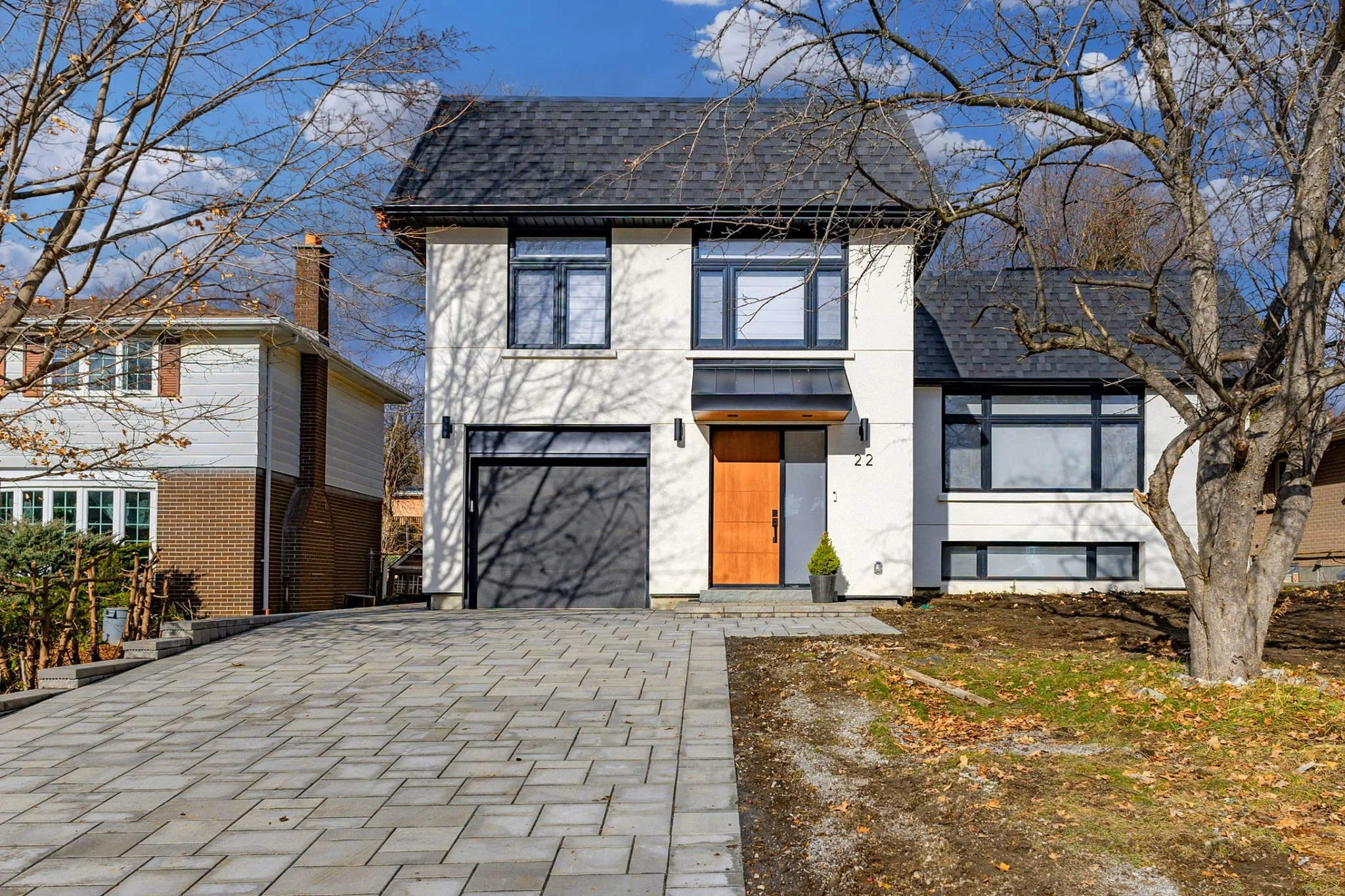Modern two-story house with white exterior, large windows, gray roof, and a wooden front door, surrounded by a driveway, trees, and a small lawn.