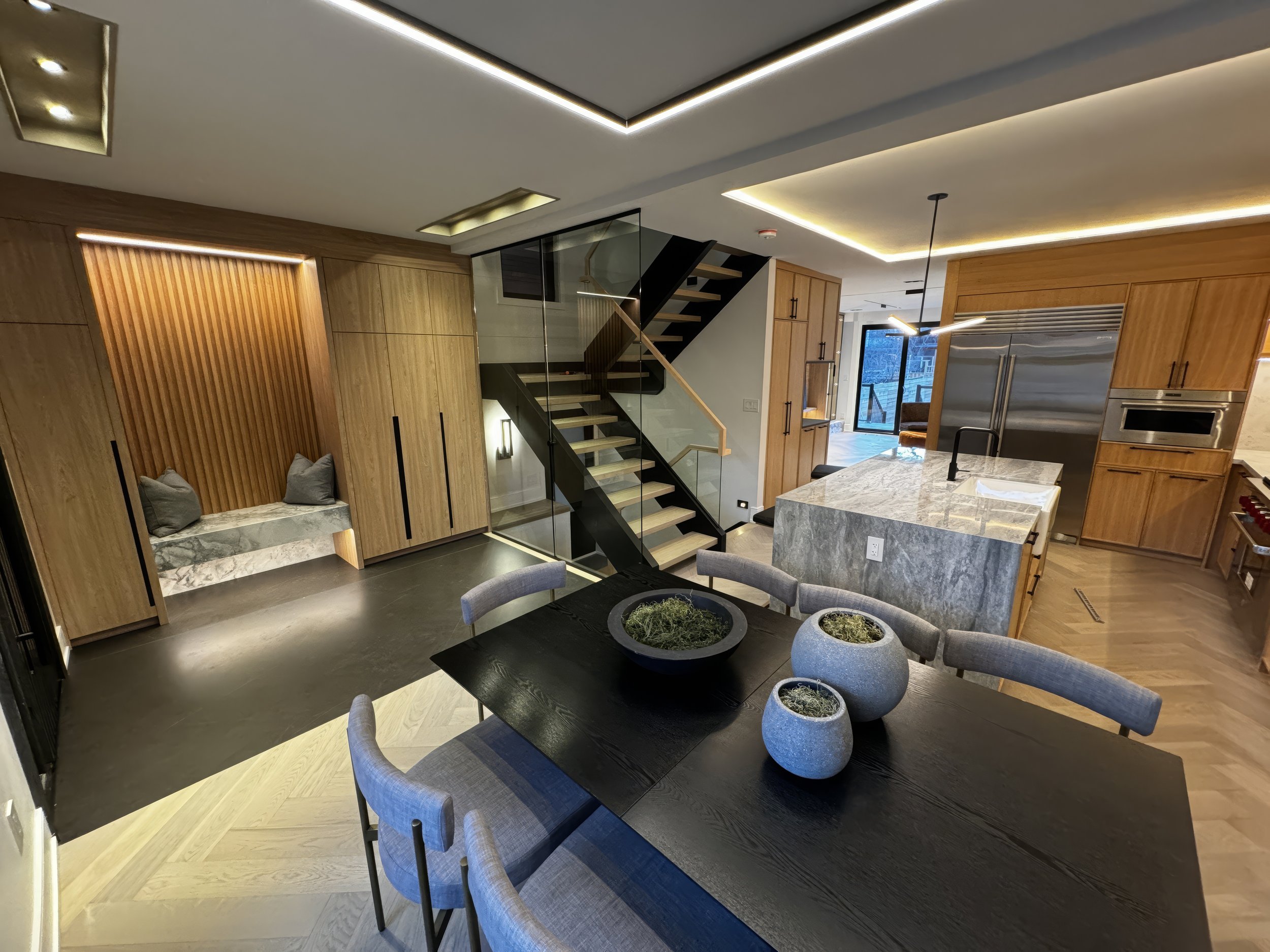Modern dining and kitchen open-concept space with marble island, wood cabinetry, floating staircase with glass railing, and built-in entryway seating.