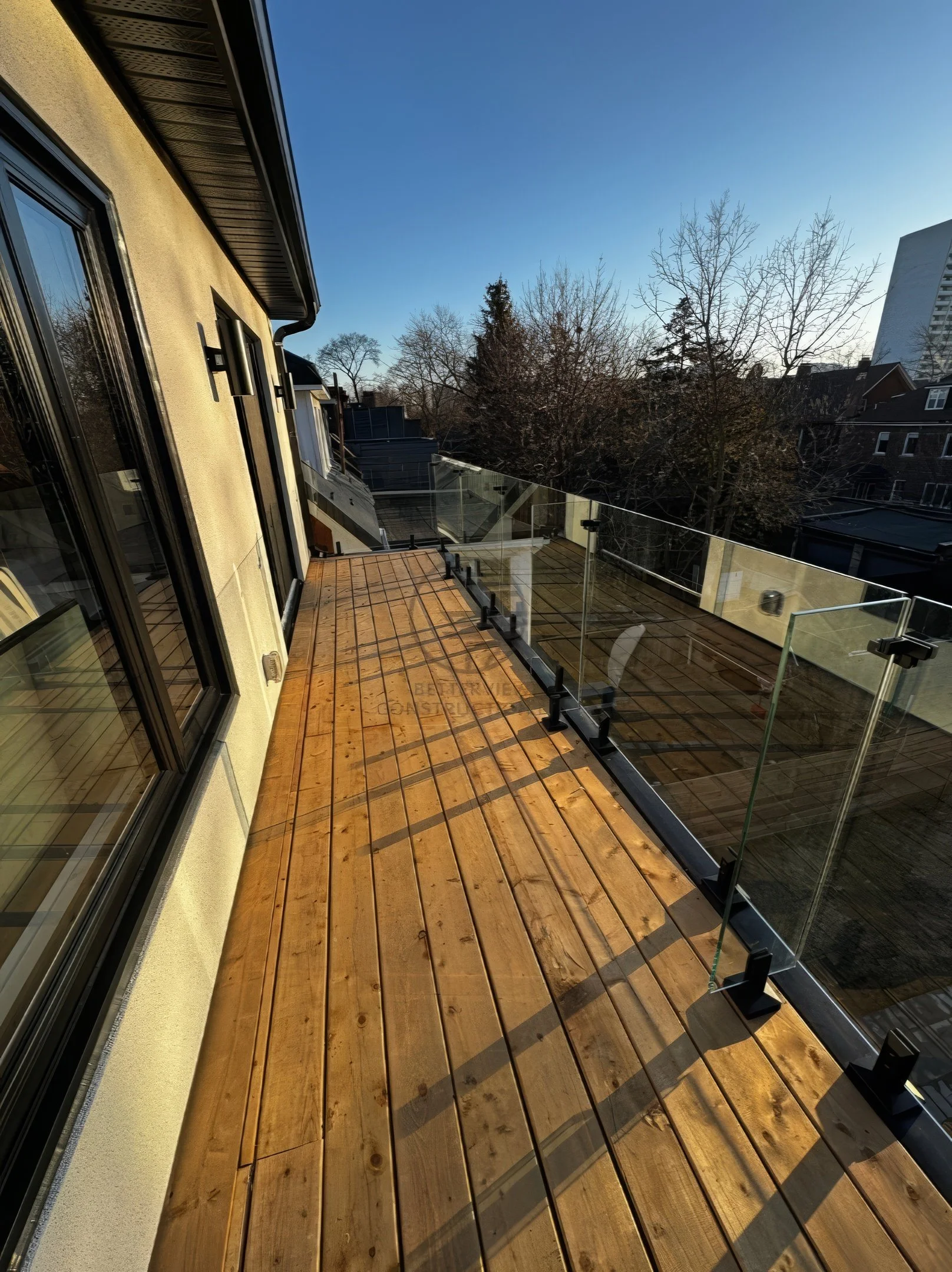 Modern balcony with wood deck flooring, glass railing system, exterior wall, and scenic outdoor neighborhood view in contemporary home design.