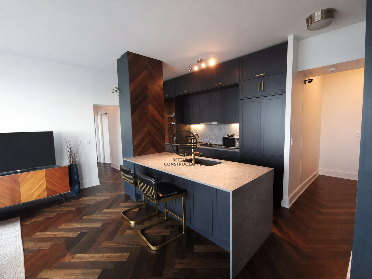Modern kitchen with dark cabinetry, large island, pendant lighting, and herringbone wood flooring in an open-concept living space.