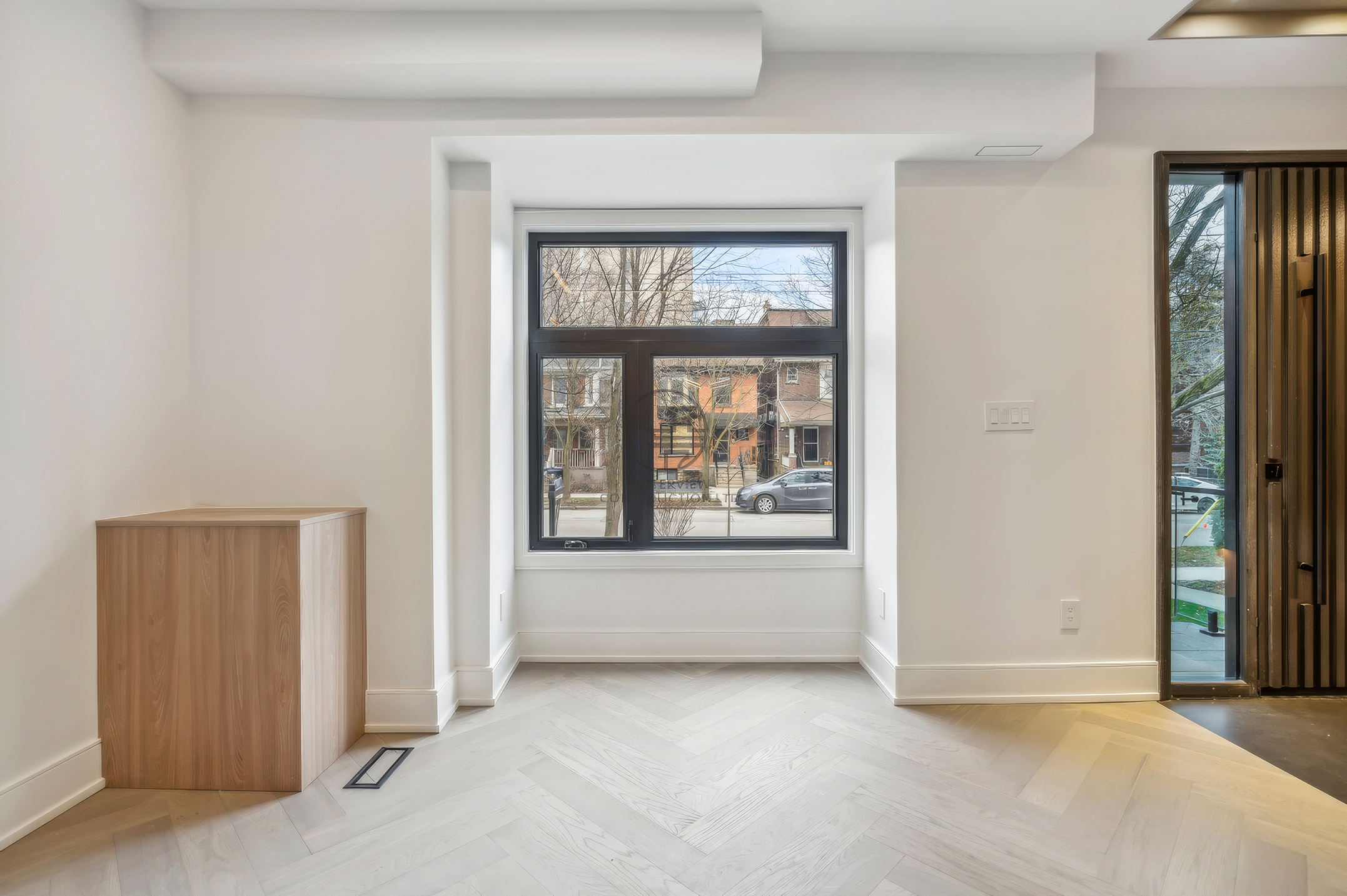 Bright minimalist interior with herringbone wood flooring, large window, clean white walls, and modern entryway connection with wood accents.