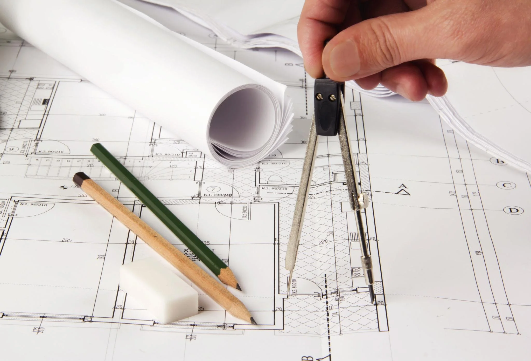 Architectural blueprints with a hand holding a pencil, a compass, and a rolled-up section of blueprint on top.