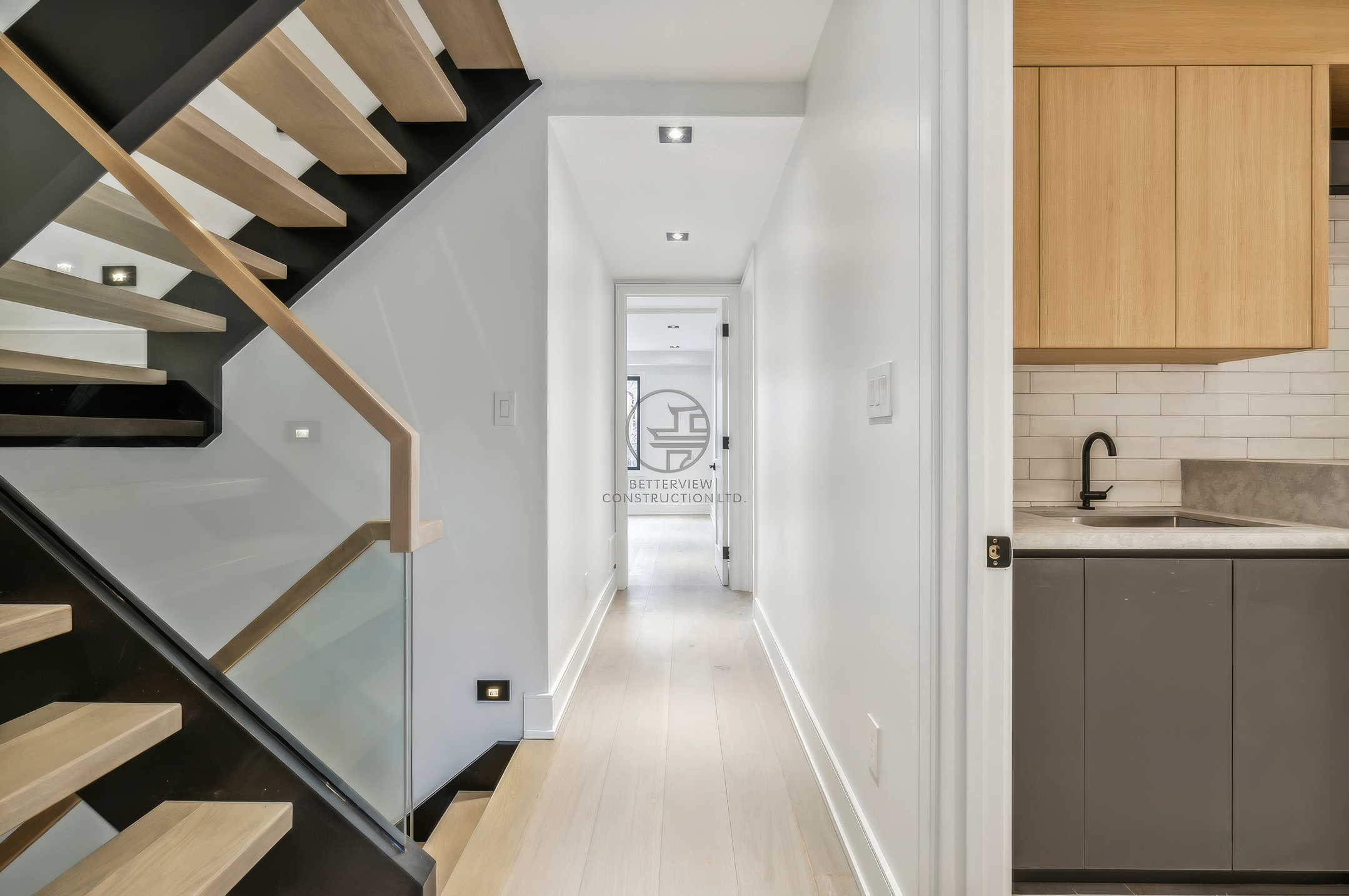 Modern hallway with floating staircase, glass railing, light wood flooring, and adjacent kitchen area with wood cabinetry and minimalist design.