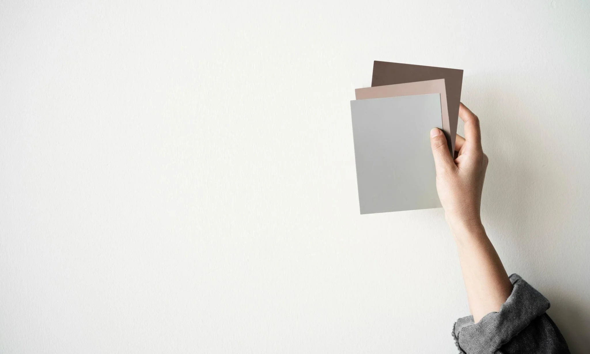 A person's hand holding three color swatches or samples against a plain white wall.