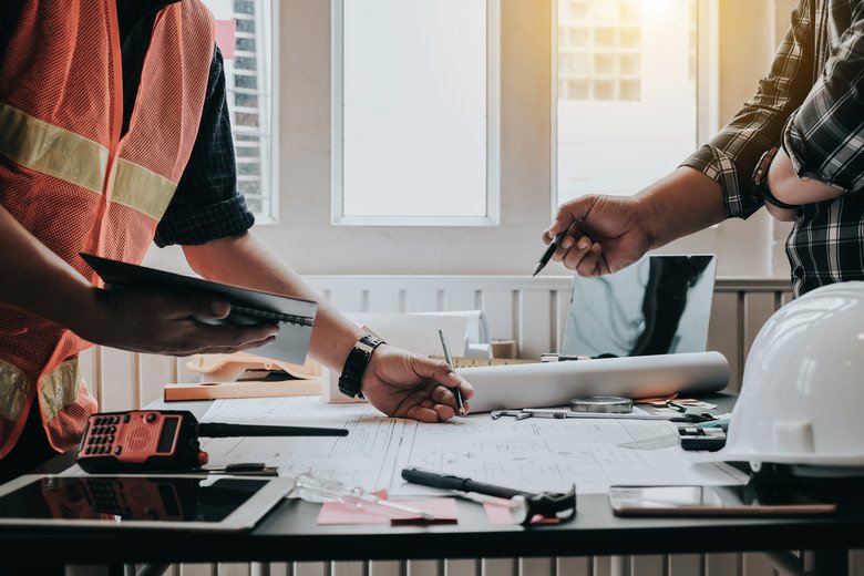 Two people discussing construction plans at a desk with blueprints, a safety helmet, and tools.