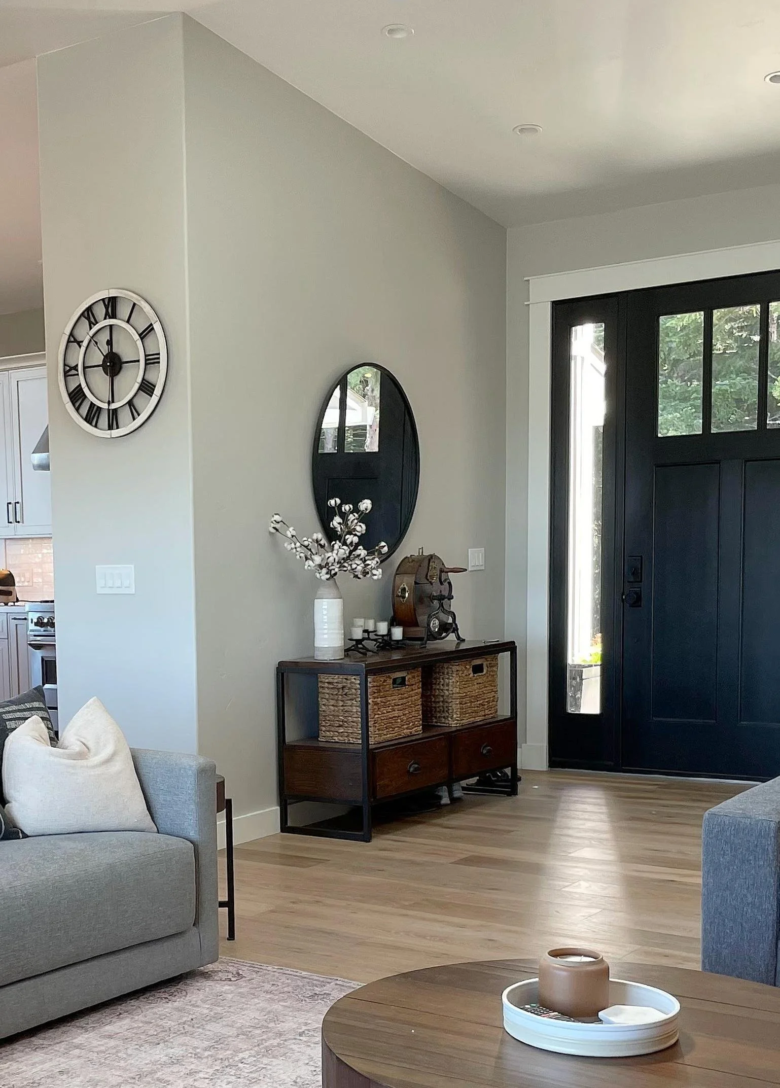 Living room with gray sofa, round wooden coffee table with a tray, a gray and beige area rug, a console table with wicker baskets, a large black front door with glass panels, a round mirror, clock, and a vase with decorative branches on the wall.