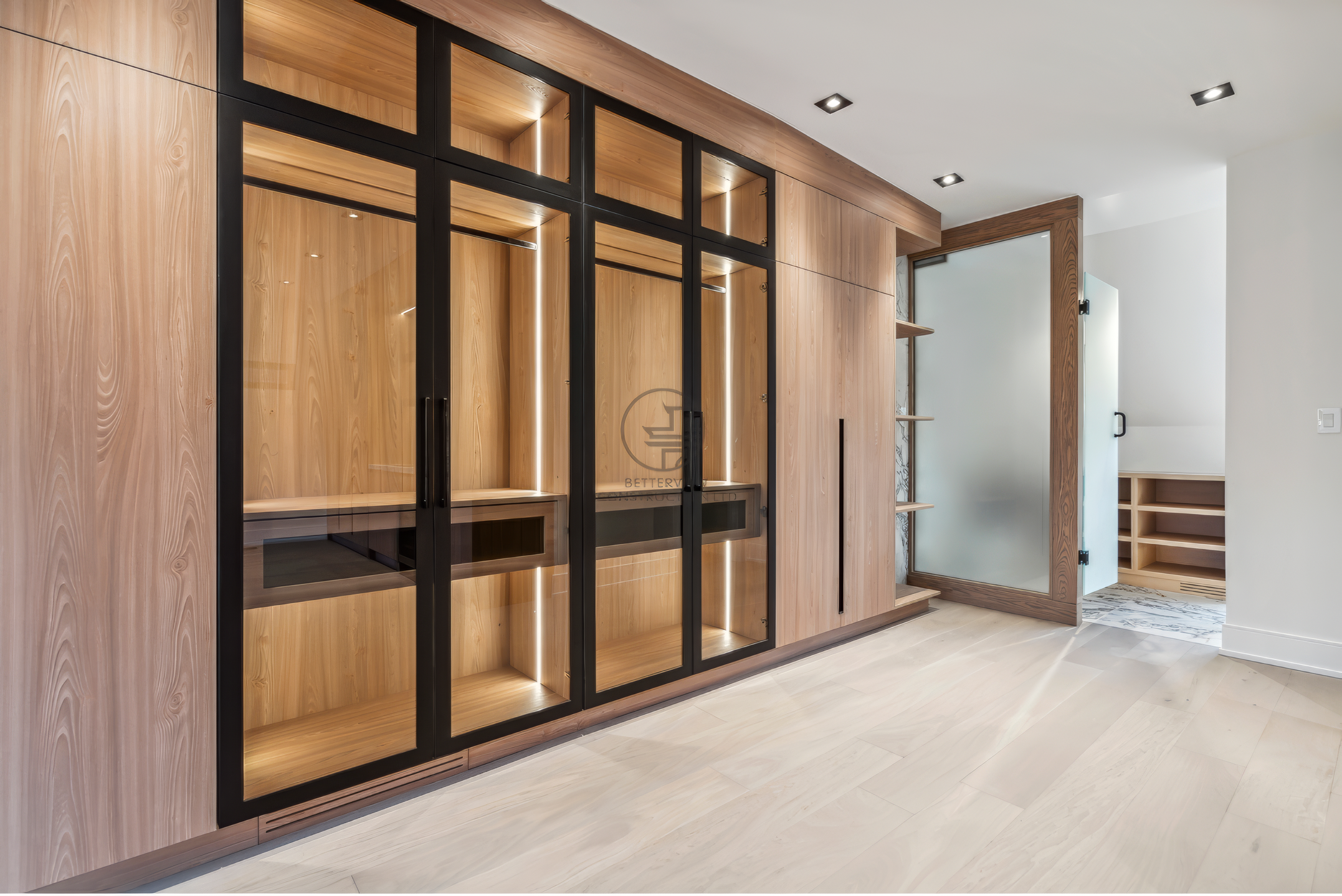Custom walk-in closet with wood cabinetry, glass doors, built-in lighting, and shelving in a modern luxury home renovation project