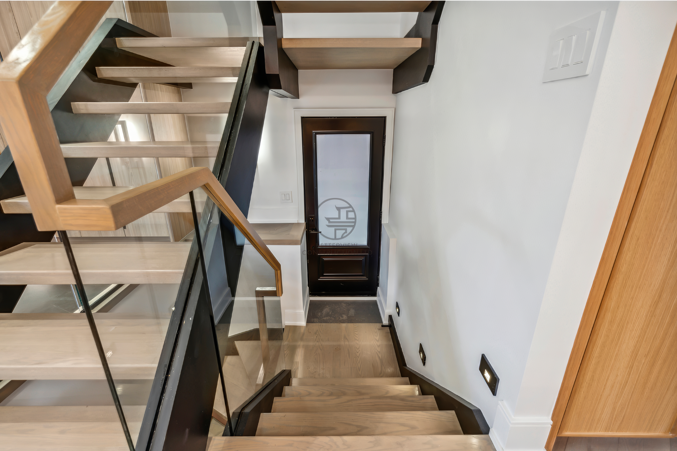 Modern staircase with glass railing, wood steps, and black steel structure leading to lower level in contemporary home interior.