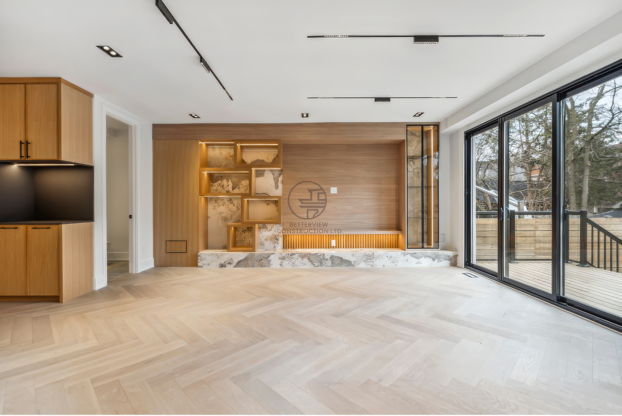 Spacious modern living room with custom wood feature wall, built-in shelving, marble base, herringbone flooring, and large sliding glass doors in a luxury home renovation project