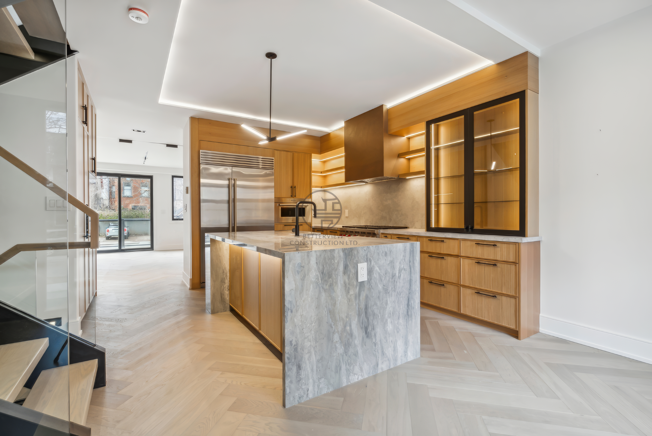 Modern open-plan kitchen with marble island, wood cabinetry, LED ceiling lighting, glass display cabinets, and herringbone wood flooring.