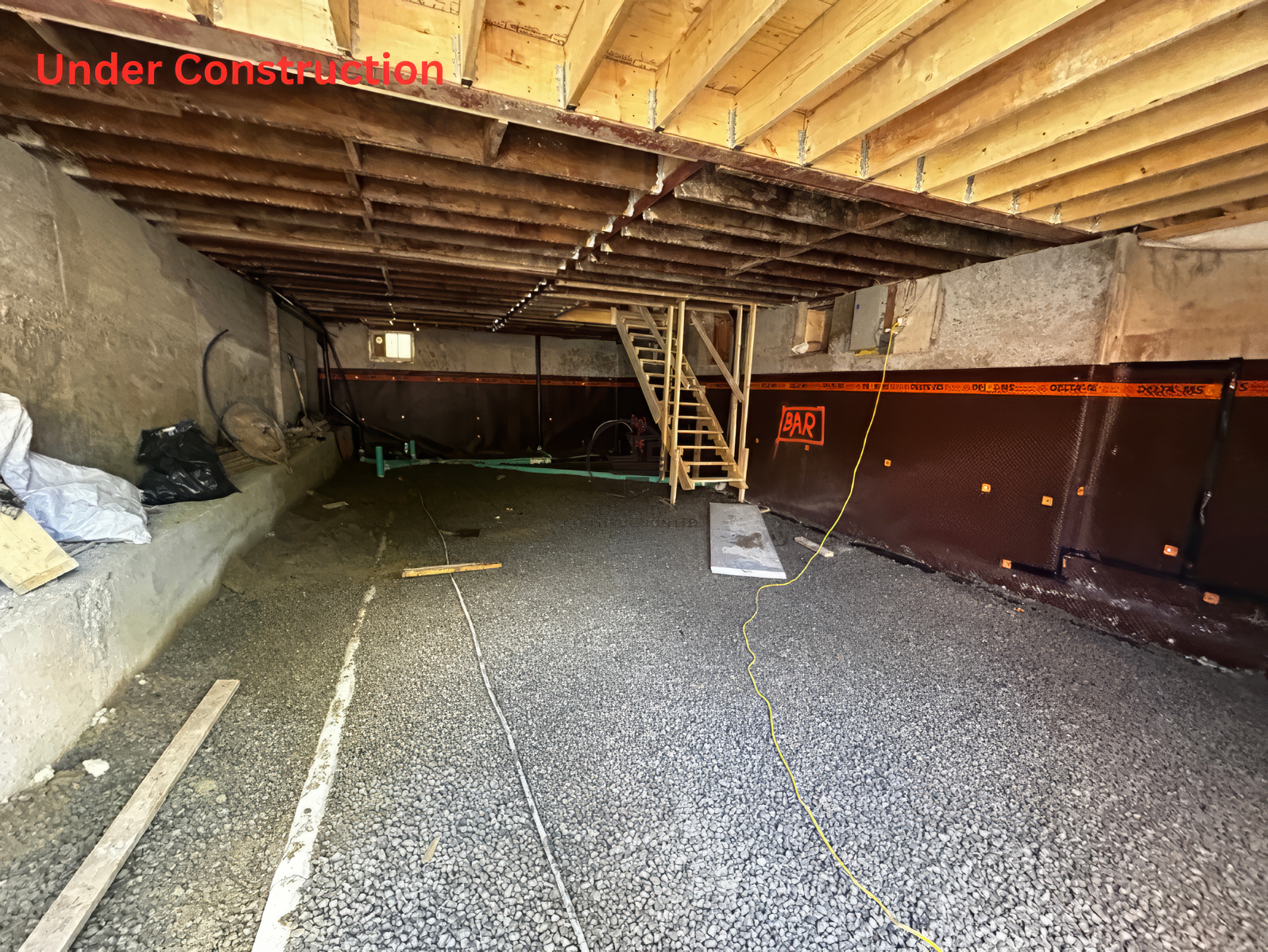 Under construction basement with gravel subfloor, exposed ceiling joists, waterproofed foundation walls, and staircase access in a residential renovation project