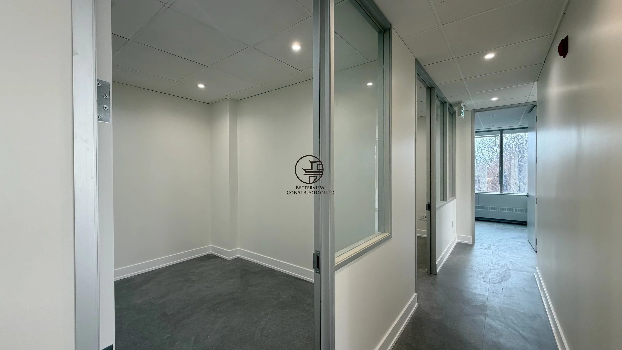 Modern office hallway with interior glass window, private rooms, and recessed lighting in commercial renovation