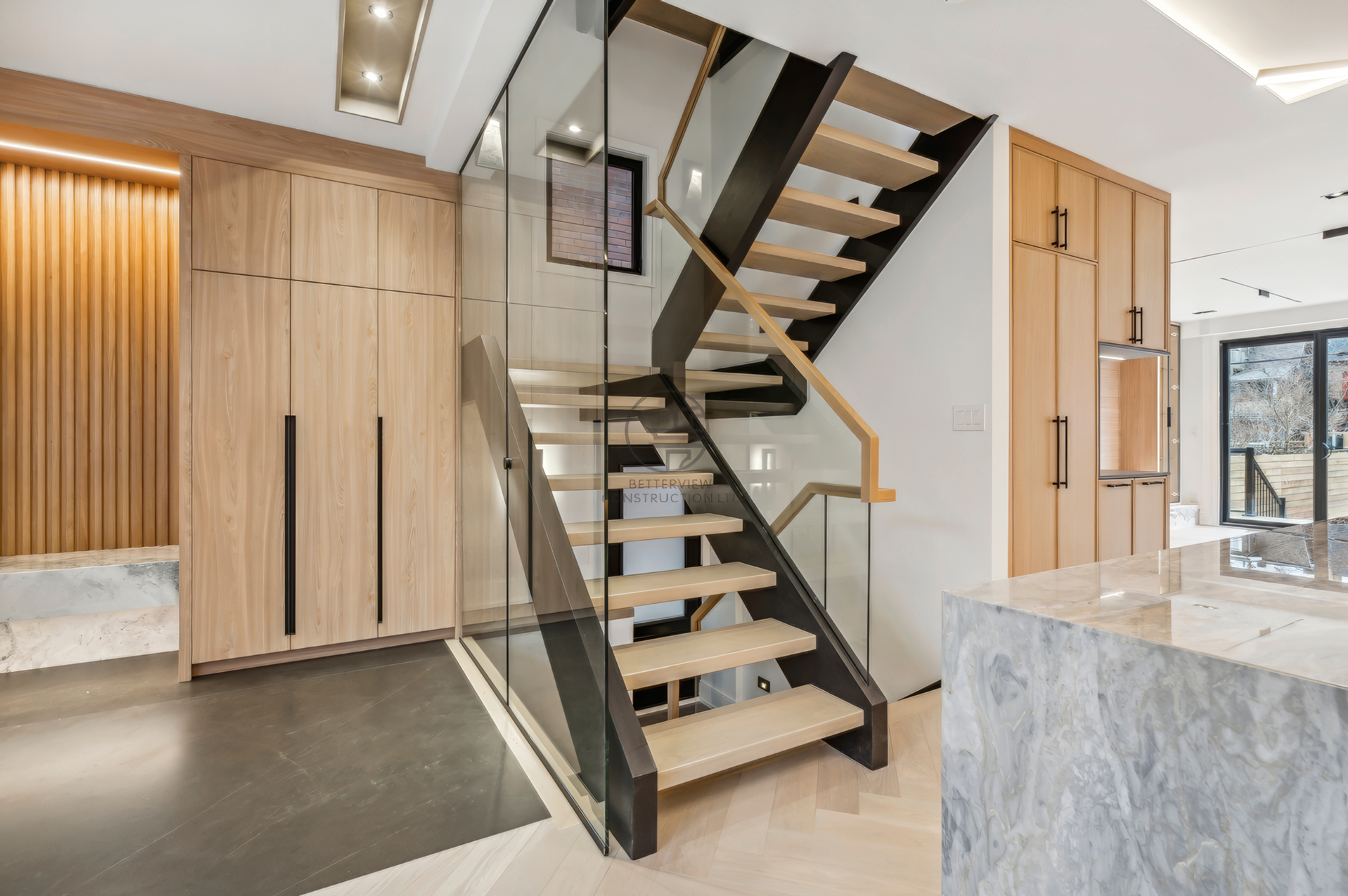 Modern open-concept interior with floating staircase, glass railing, wood cabinetry, and marble kitchen island in contemporary home design.