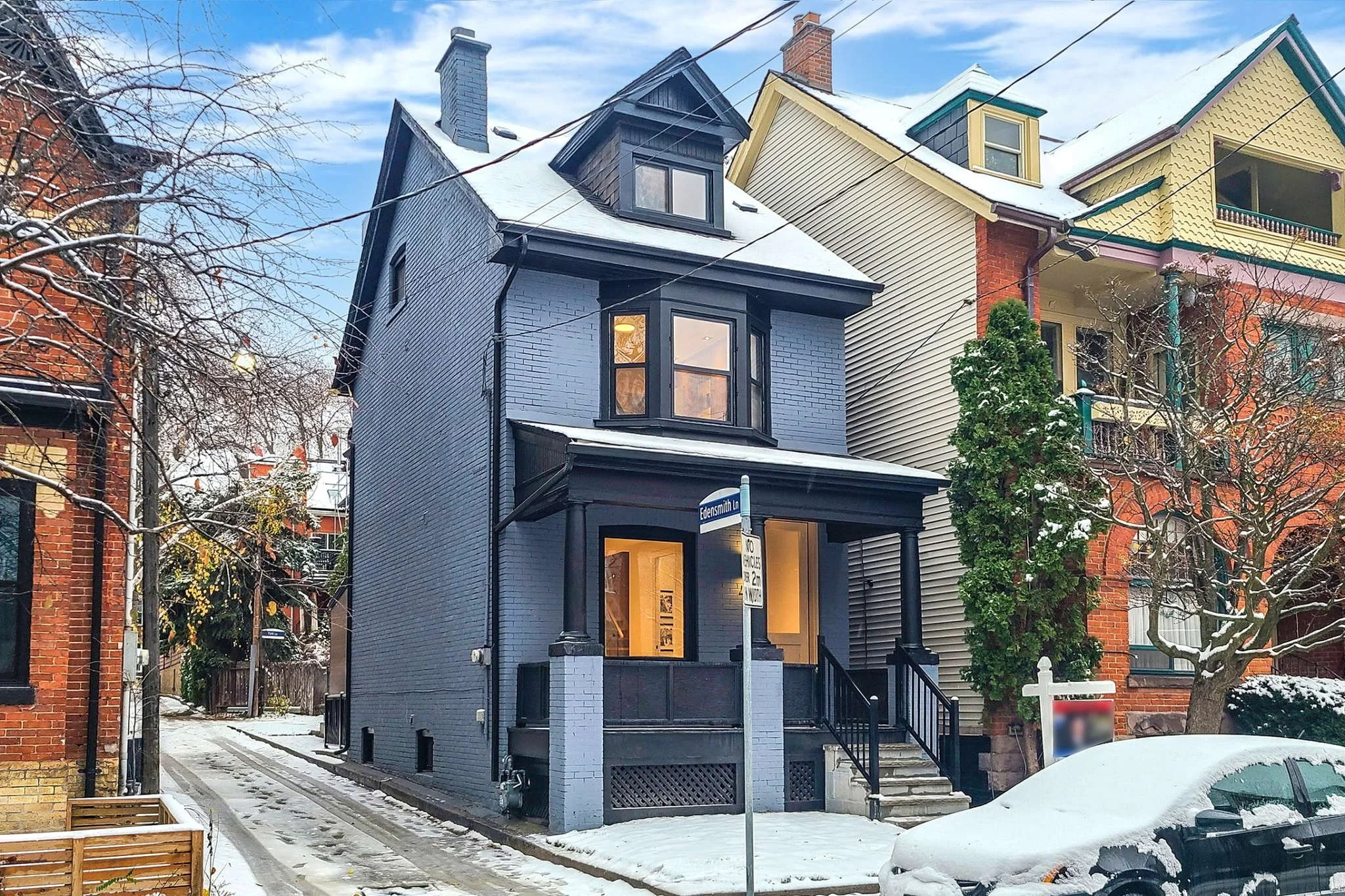 A blue-painted, three-story Victorian-style house with a small porch, bay windows, and a snow-covered roof in an urban neighborhood during winter, with neighboring houses, trees, and a street sign visible.