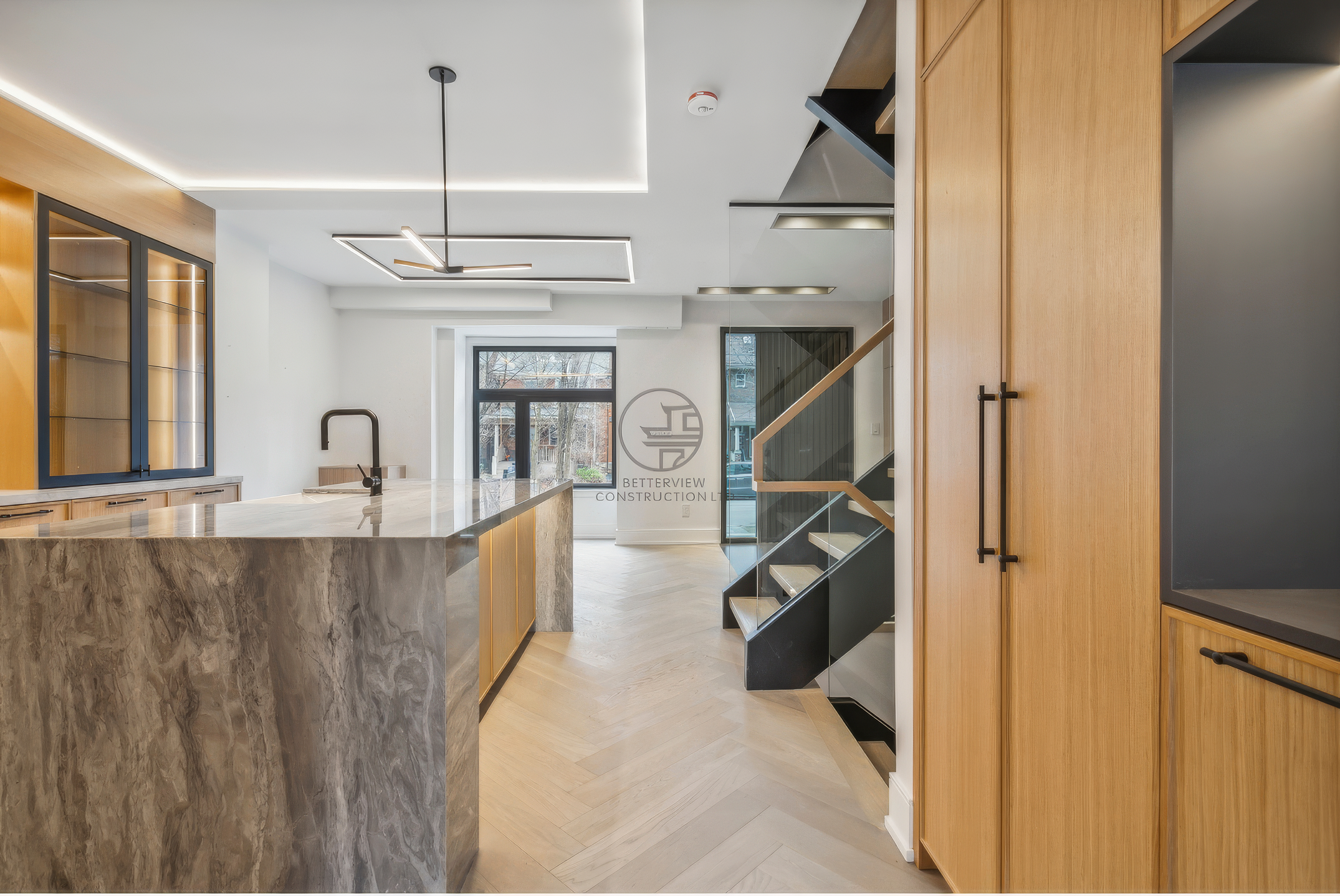 Modern kitchen with marble island, wood cabinetry, open layout, LED ceiling lighting, and adjacent floating staircase with glass railing.
