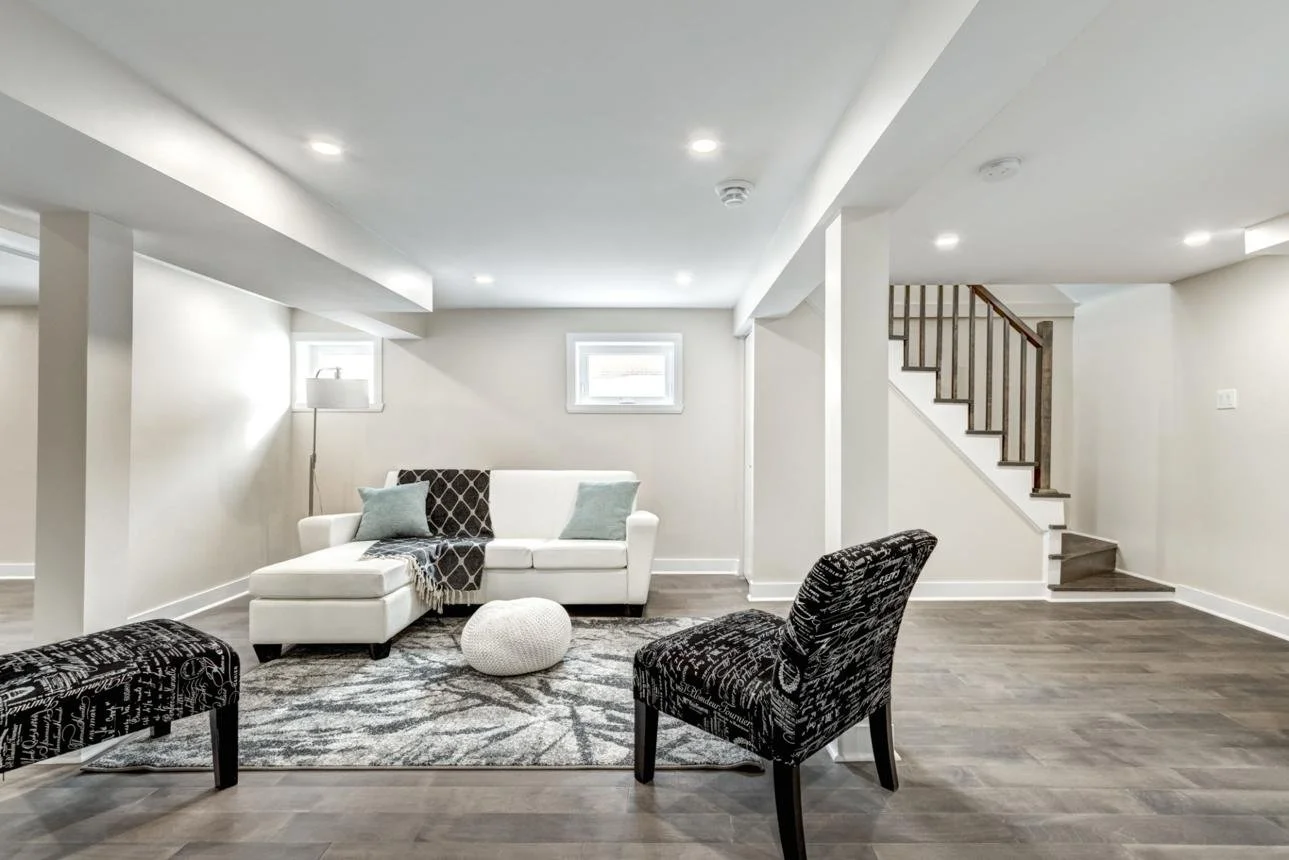 Living room with white walls, wood flooring, a white sofa with pillows, two patterned chairs, a patterned rug, a floor lamp, two small windows, and a staircase with wooden railing.