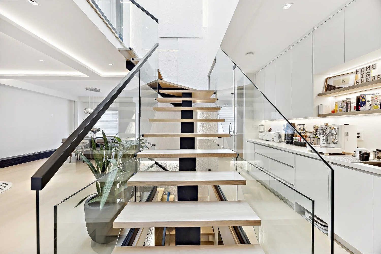 Modern staircase with wooden steps and black support, glass railing, in a bright open kitchen and living space with white walls and cabinetry, plants, and shelves with kitchen appliances and decorations.