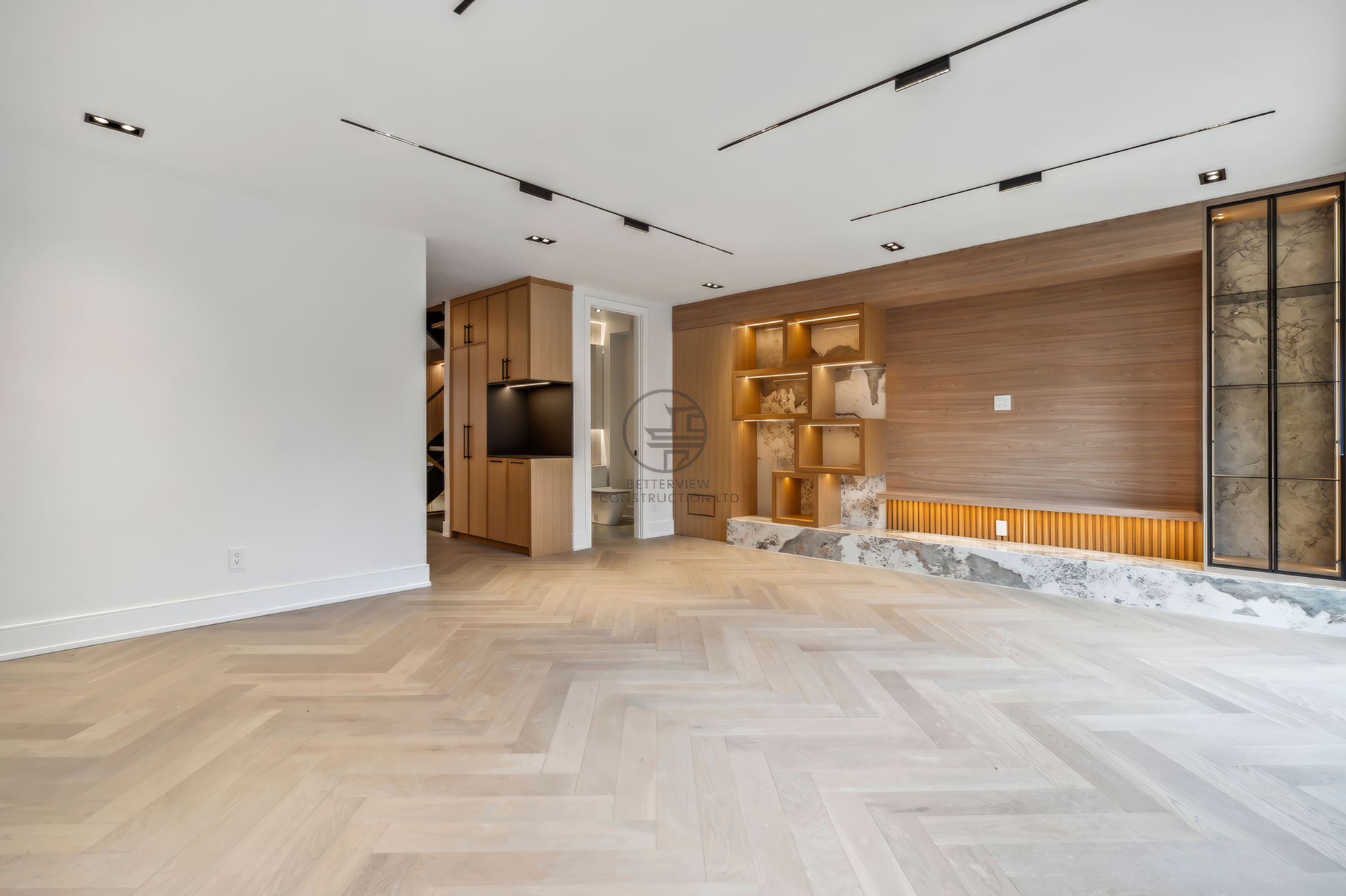Open-concept living room with herringbone wood flooring, wood feature wall with built-in shelves, recessed lighting, and modern minimalist design.