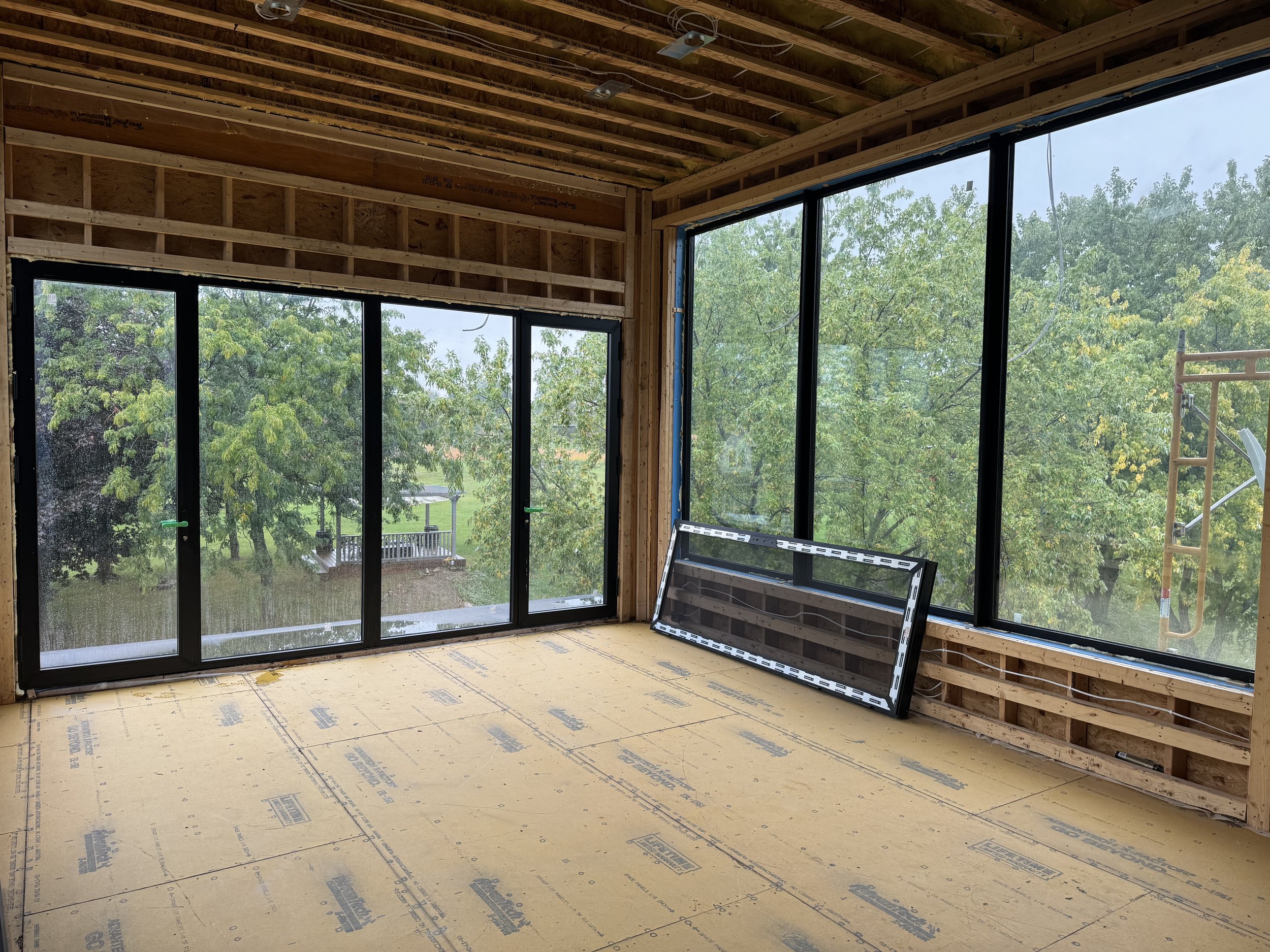 Interior of a building under construction with framed wooden walls and large windows, showing trees outside.