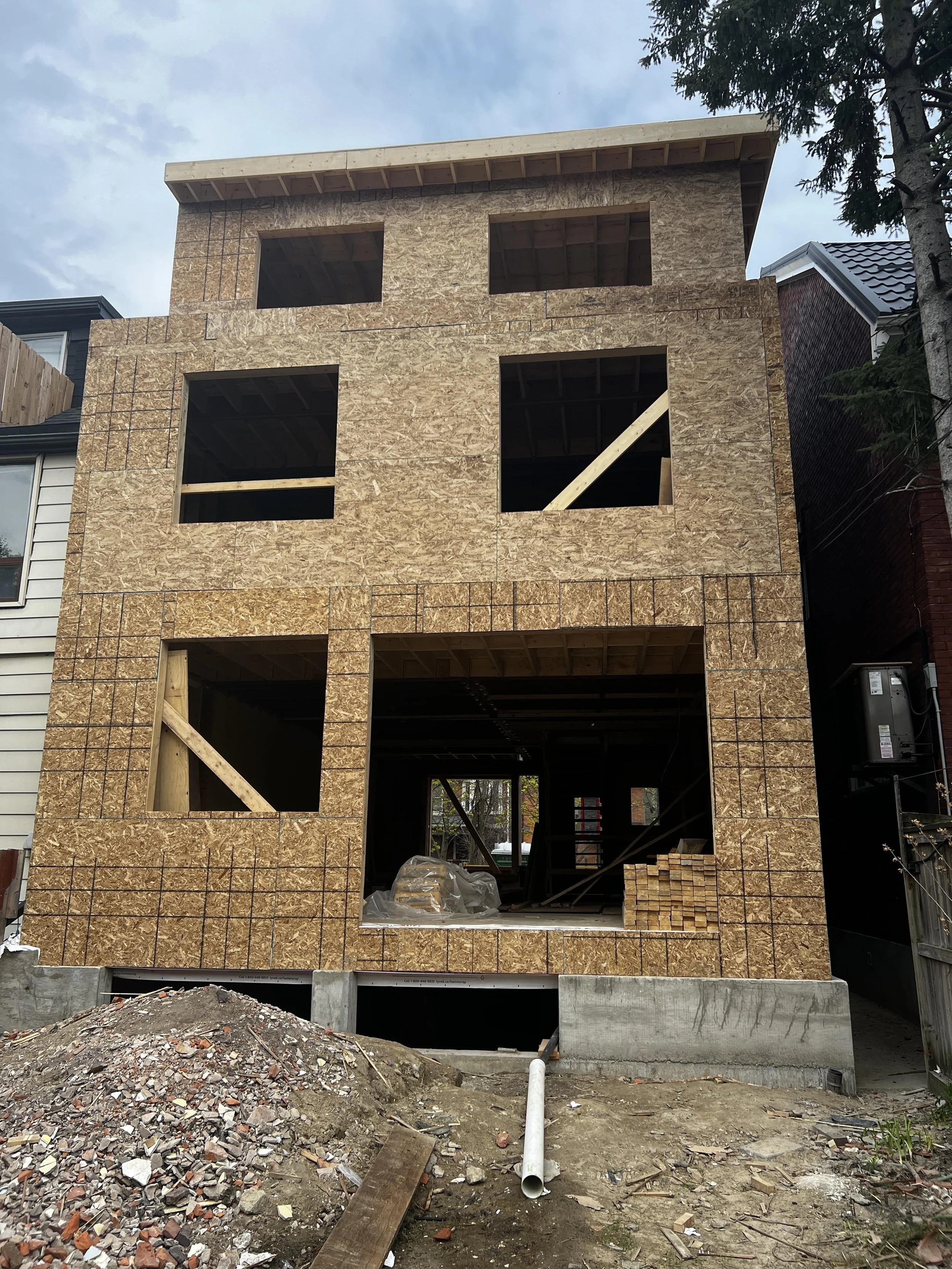 A multi-story house under construction with exposed plywood walls and open window frames, surrounded by construction debris and materials, with neighboring houses and trees in the background.