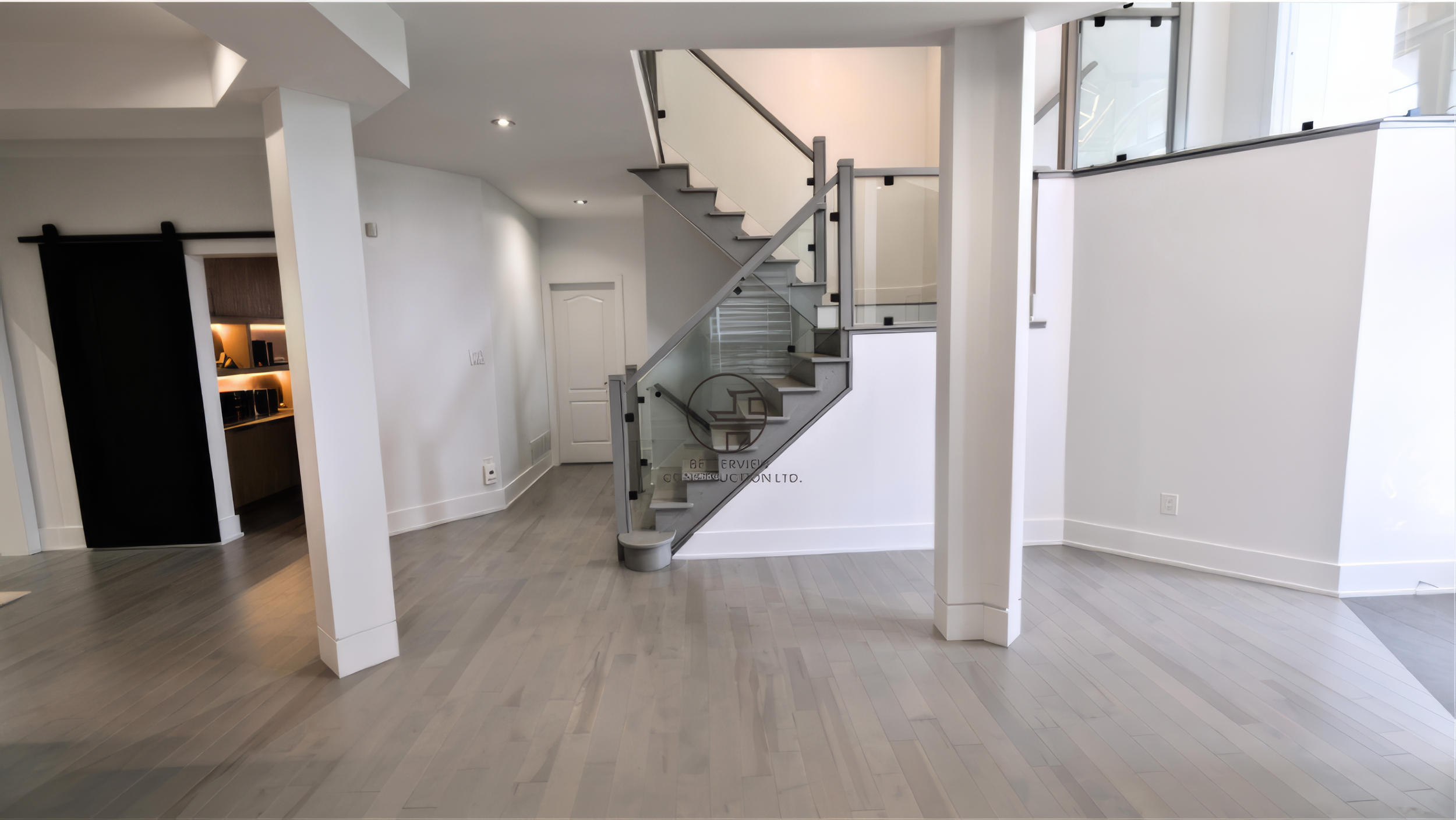 Open concept basement renovation featuring a modern glass staircase, light wood flooring, and spacious minimalist layout with structural columns and bright interior lighting