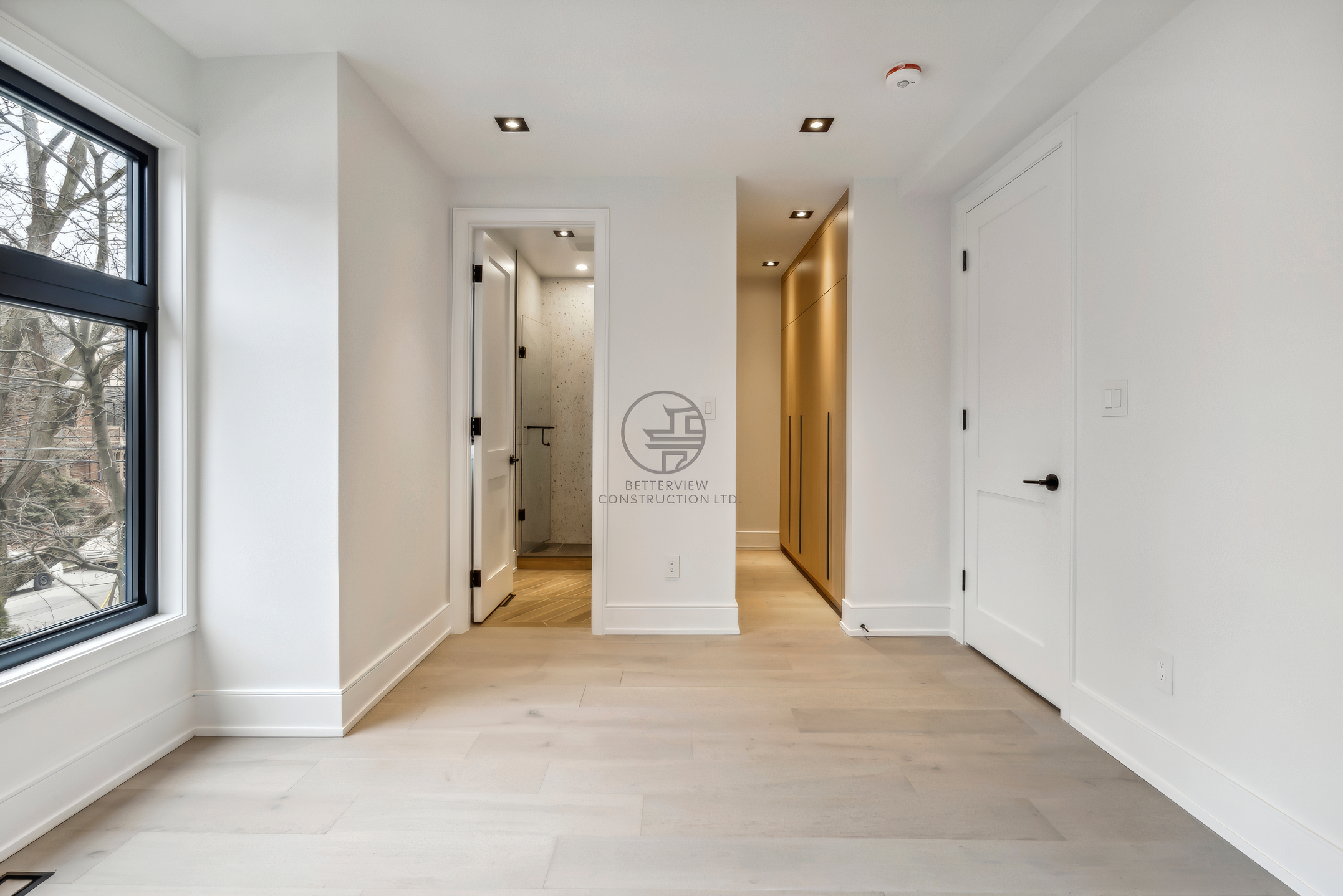 Modern hallway interior with light wood flooring, built-in closet storage, recessed lighting, and clean white walls leading to bathroom space.