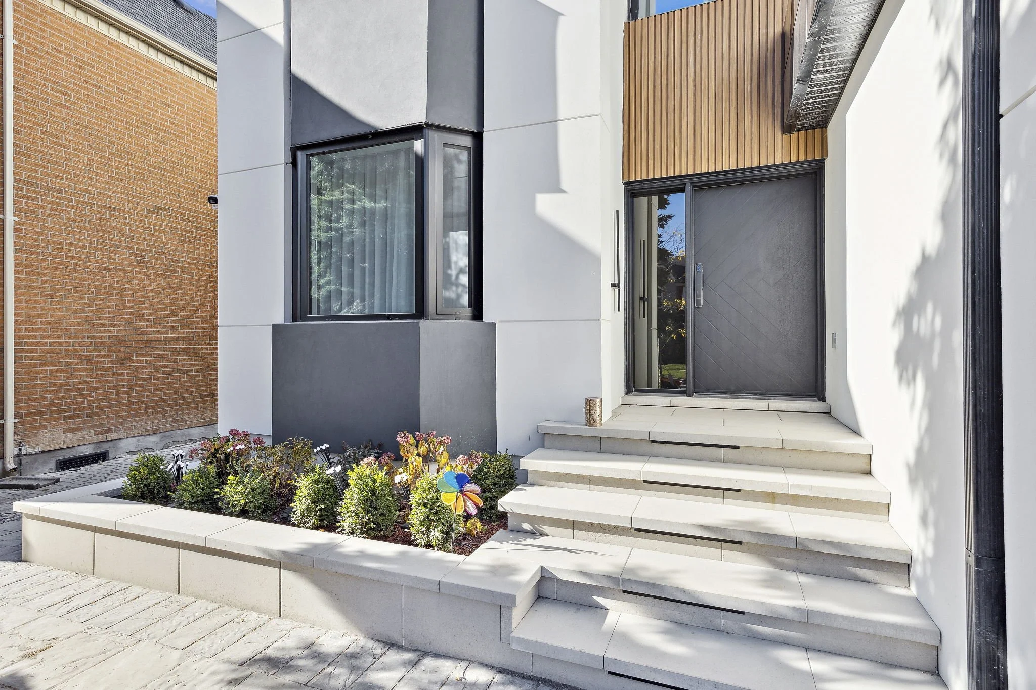 Modern house entrance with white steps leading to a black door, surrounded by a flower bed with colorful flowers, a brick and white wall, and a wooden accent on the house facade.