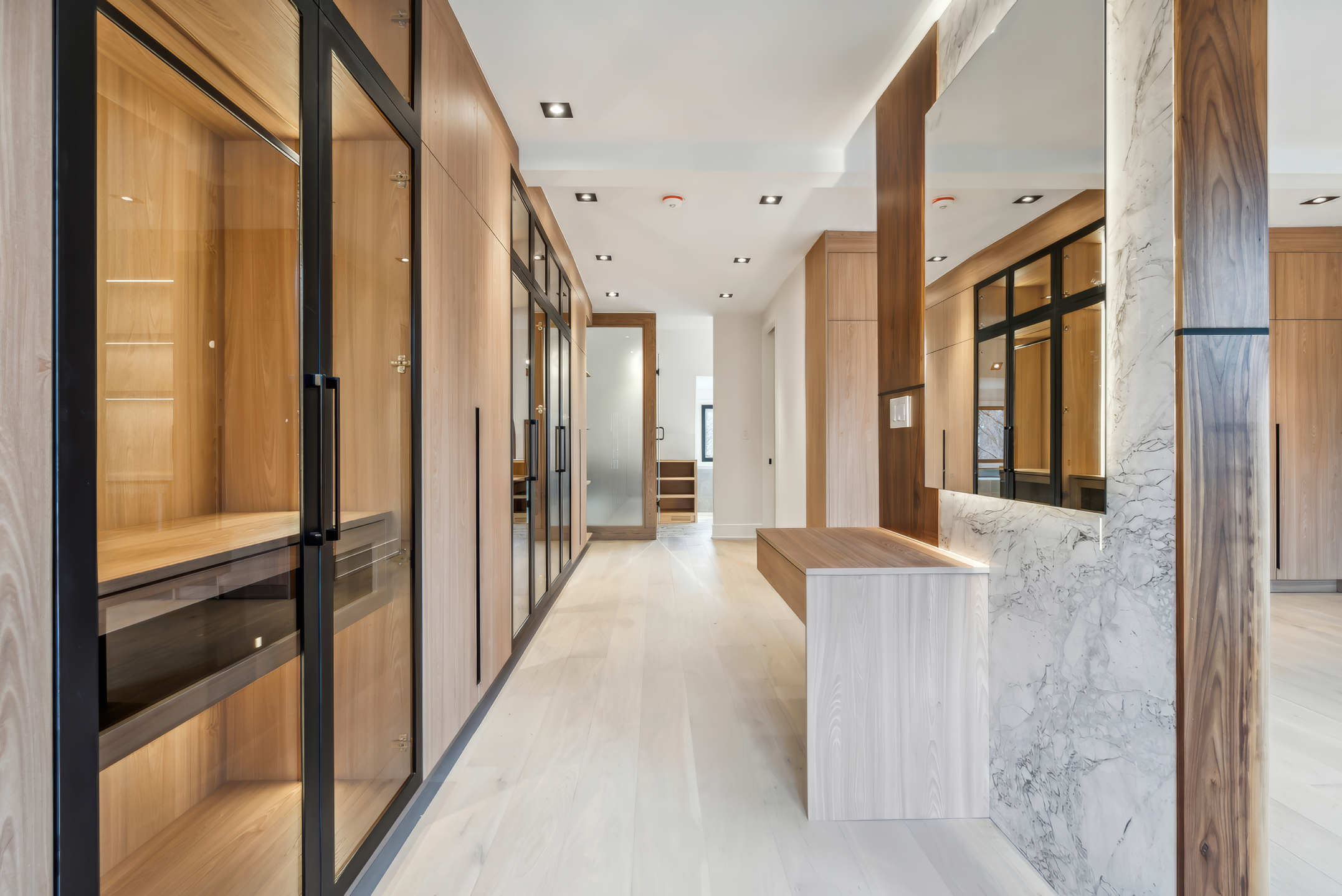 Modern interior hallway with custom wood cabinetry, glass display closet, marble accent column, and built-in bench in a luxury home renovation project