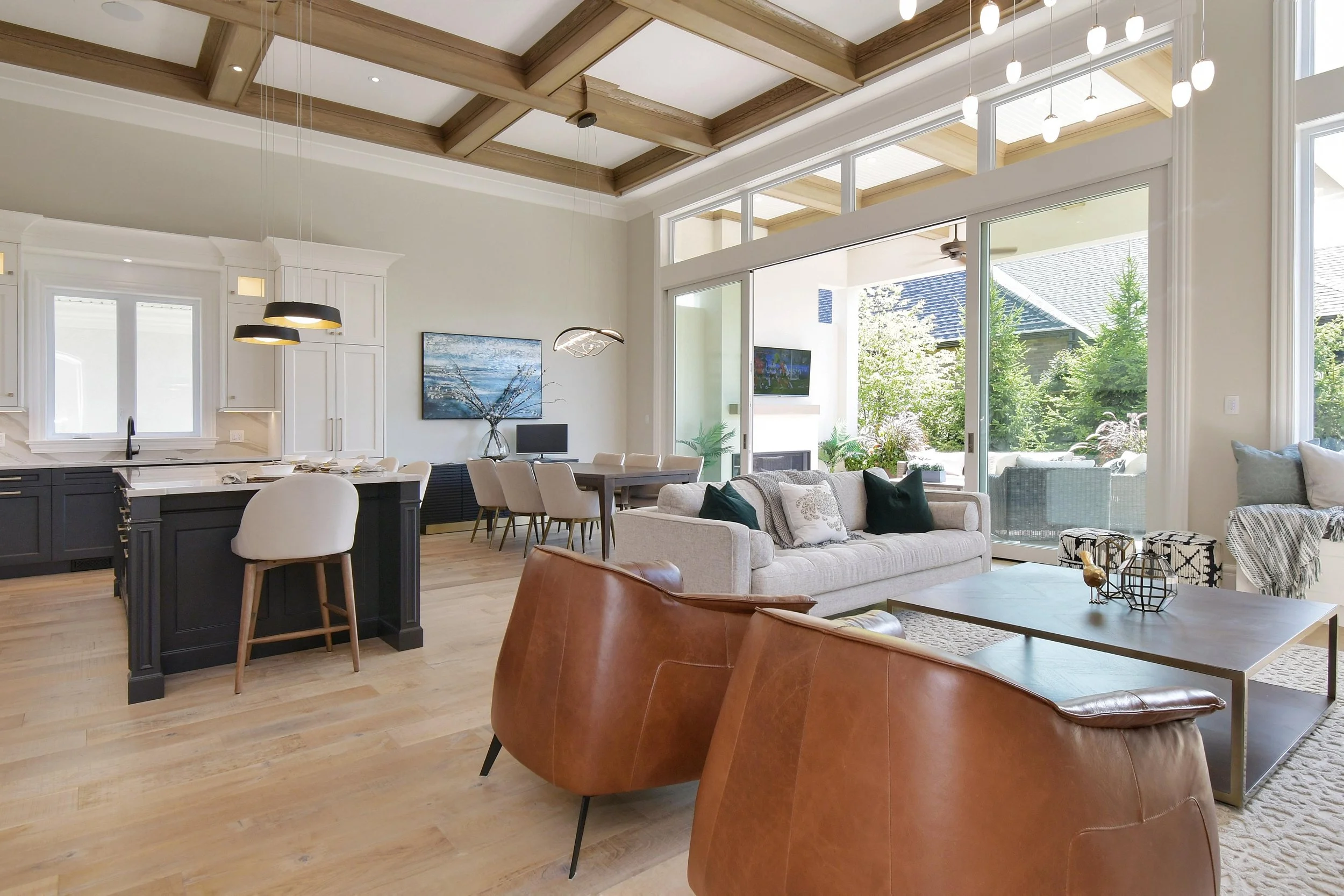 Open-concept living room and kitchen with large windows, wooden ceiling beams, a beige sofa, a brown leather chair, modern pendant lights, and outdoor patio view with greenery.