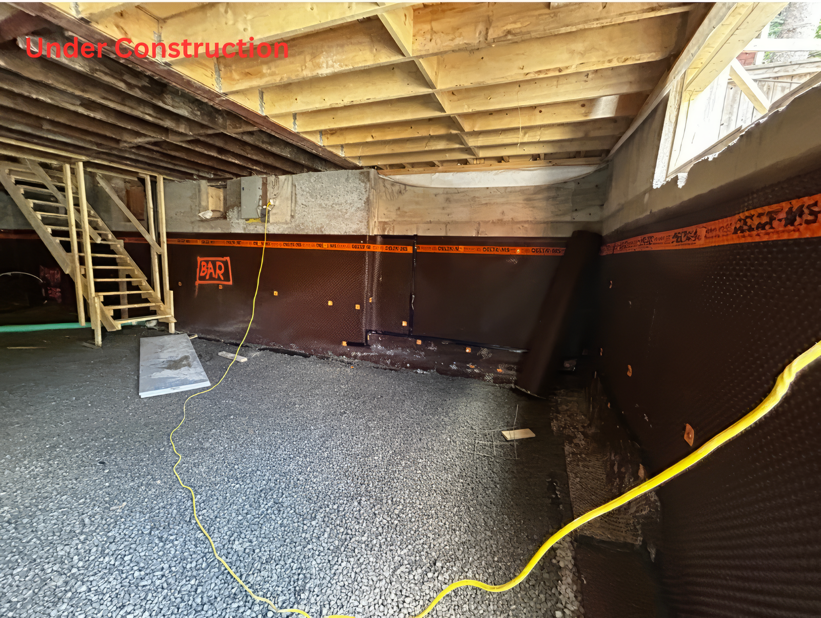 Under construction basement interior with gravel base, waterproof membrane on foundation walls, exposed ceiling framing, and staircase during home renovation project