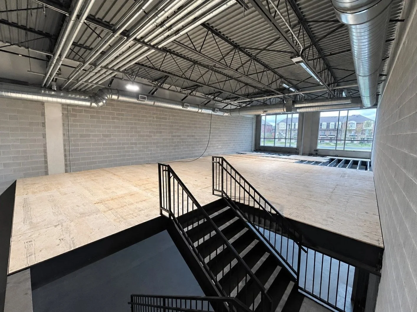 Interior of a building under construction with exposed ductwork and wiring, plywood flooring, and large windows showing houses outside.