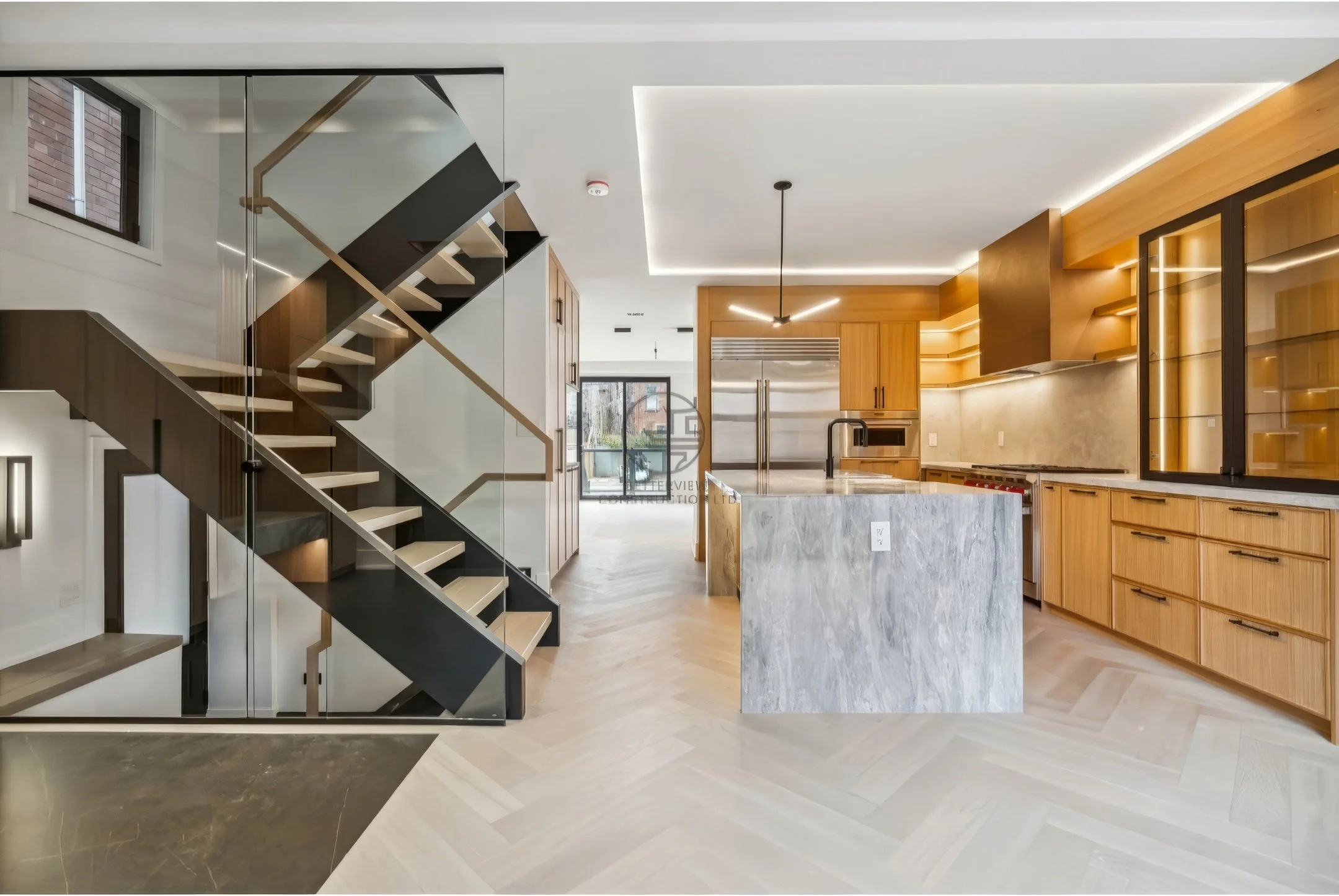 Modern open-concept kitchen with marble island, wood cabinetry, LED lighting, and floating staircase with glass railing in contemporary home design.